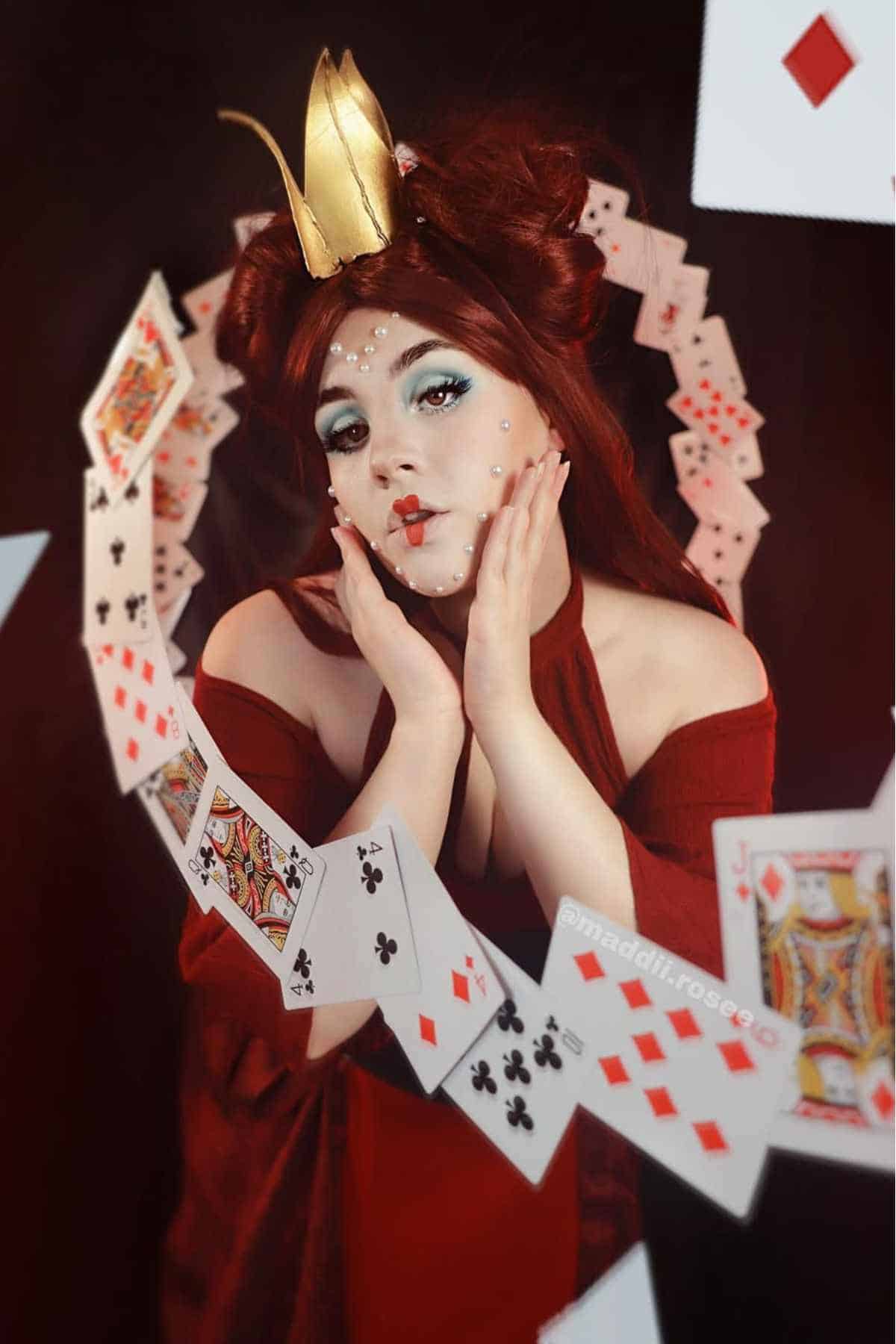 Person in a red costume and gold crown poses with hands on face, surrounded by floating playing cards. Face is decorated with white dots.