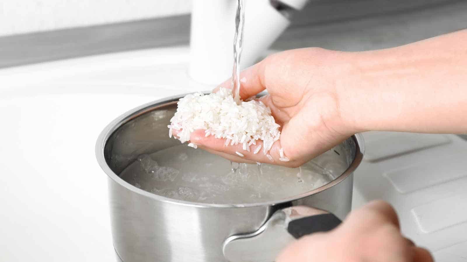 A hand rinsing white rice under running water over a stainless steel pot in a kitchen setting.