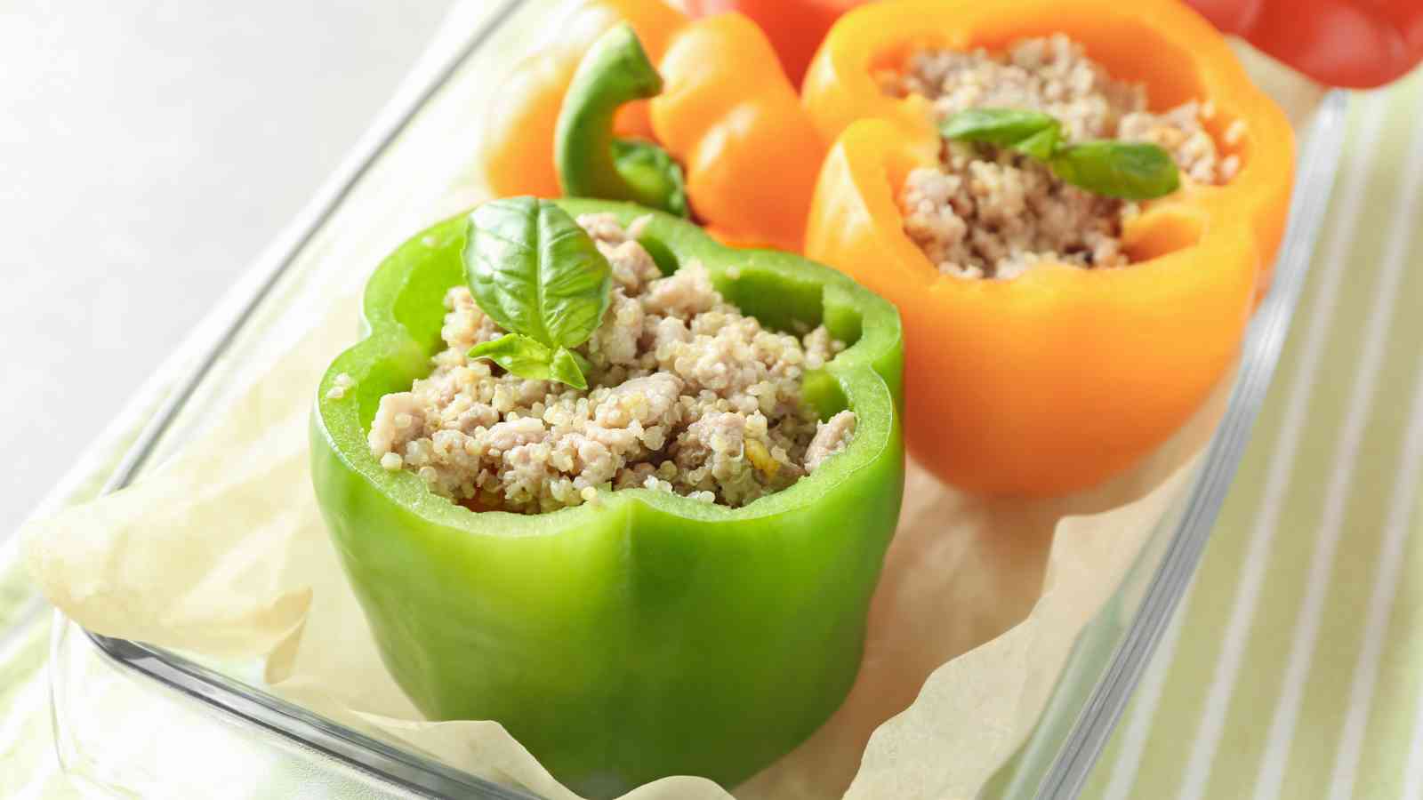 Two bell peppers, one green and one orange, filled with a mixture of grains and ground meat, topped with basil, placed in a glass baking dish.