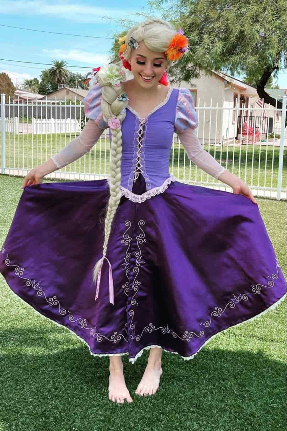 Woman in a purple princess costume with long braided blonde hair and flowers stands barefoot on grass, holding out her skirt.