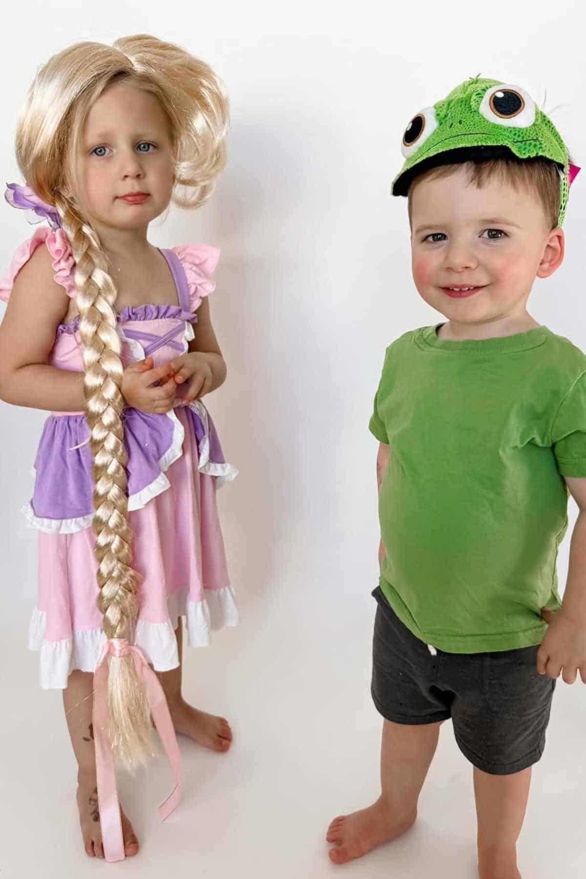 A young girl in a Rapunzel costume with a long blonde braid stands next to a boy wearing a green shirt and a chameleon hat, both against a plain white background.