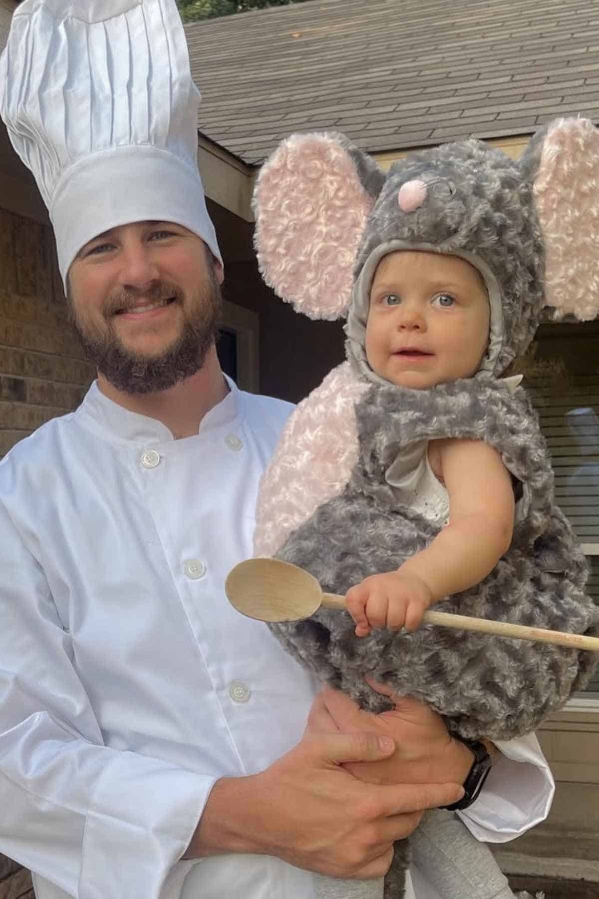An adult in a chef outfit holds a small child dressed in a gray mouse costume and holding a wooden spoon, standing outside near a house.