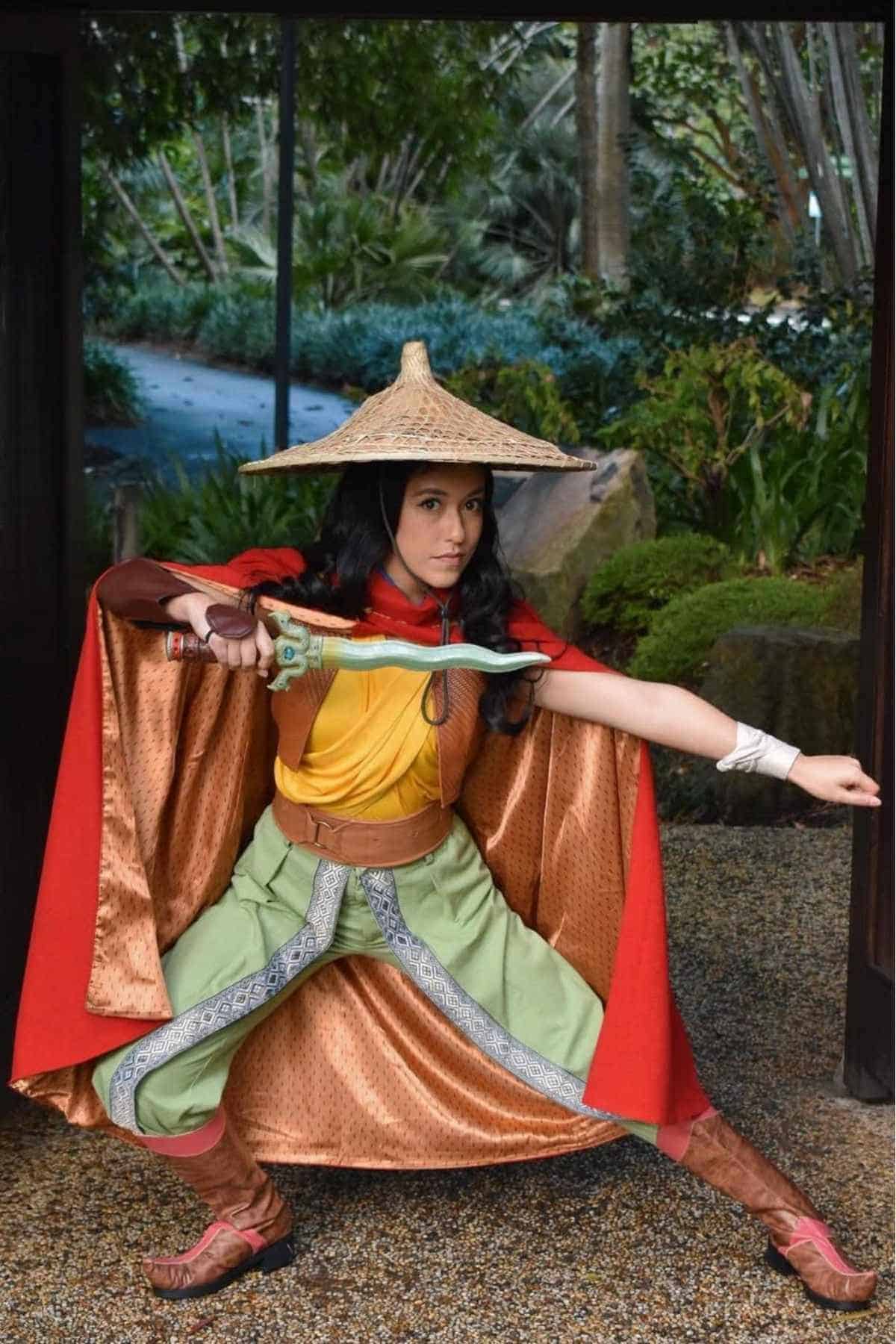Person in a detailed warrior costume with a straw hat, red cape, and sword, posing in a crouched stance in a garden setting.