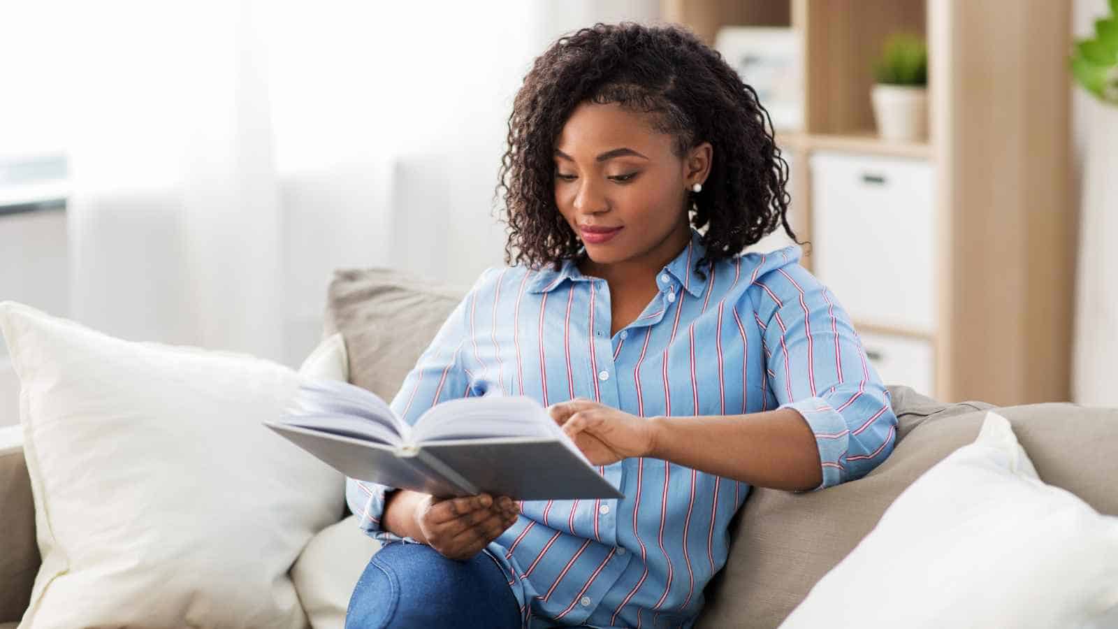 A woman sits on a couch reading a book. She wears a blue striped shirt and jeans. There are white cushions and a shelf with plants in the background.
