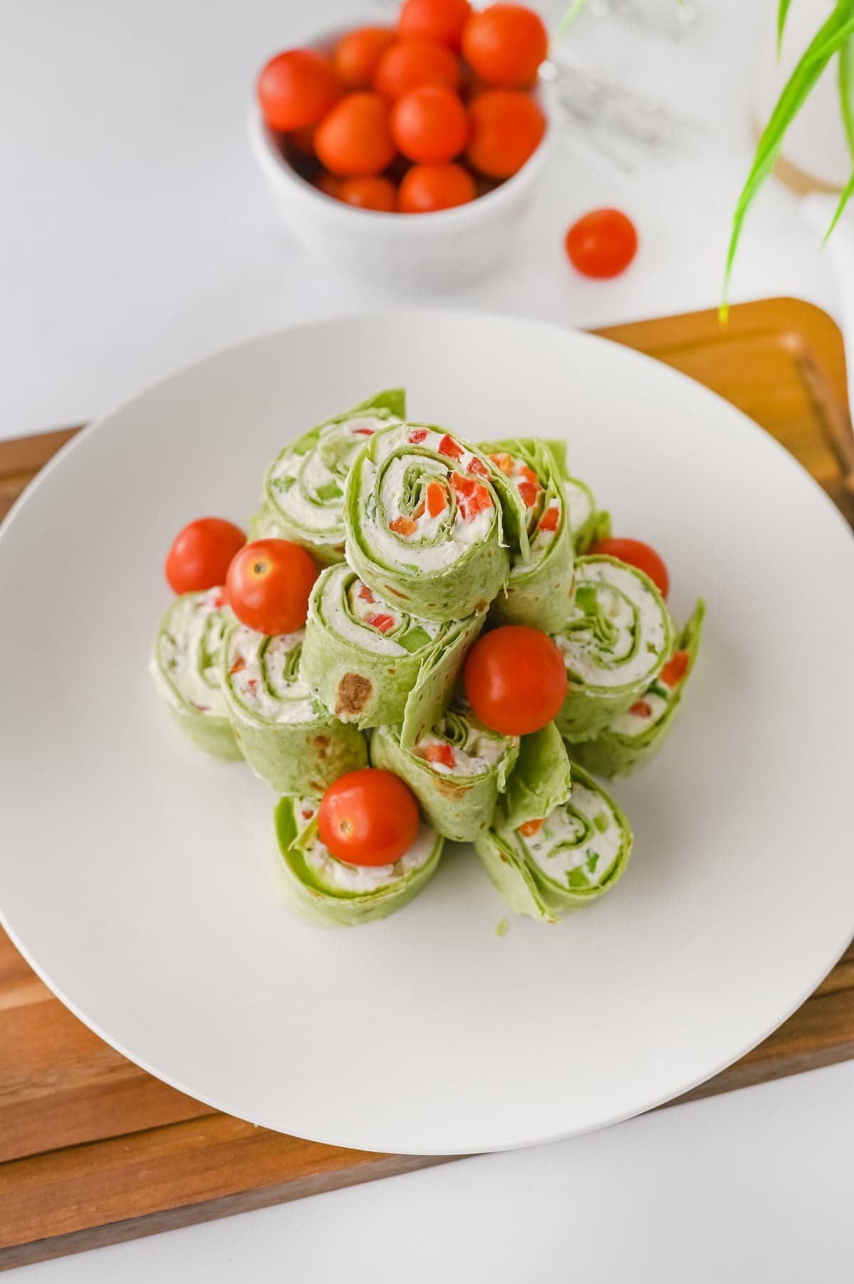A plate of spinach tortilla pinwheels filled with cream cheese and vegetables, garnished with cherry tomatoes. A bowl of cherry tomatoes is in the background.