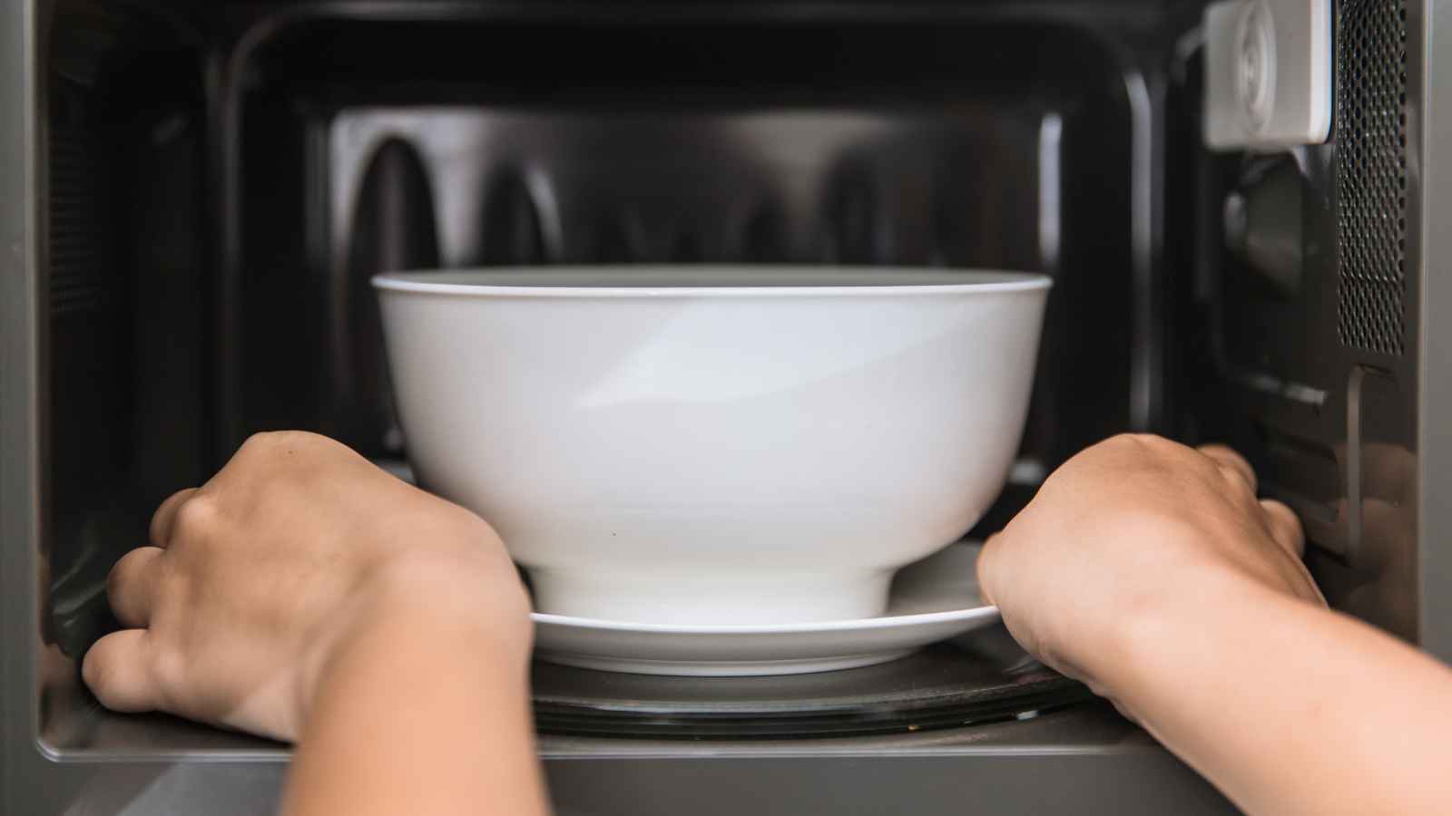A person places a white bowl on a plate inside a microwave oven.