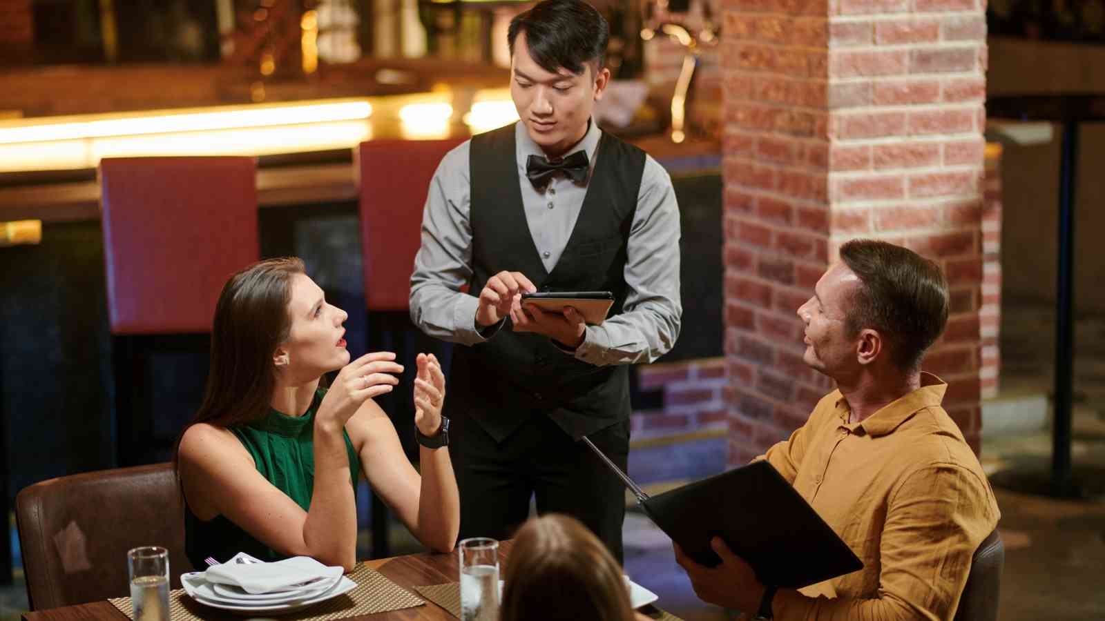 A waiter stands taking an order from two seated restaurant customers; one woman is speaking while the man looks at the menu.