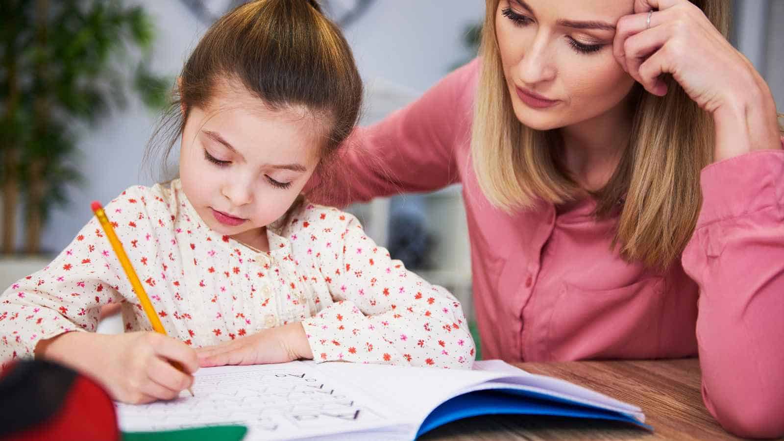 A young girl is drawing in a book with a pencil while a woman sits beside her, watching attentively.