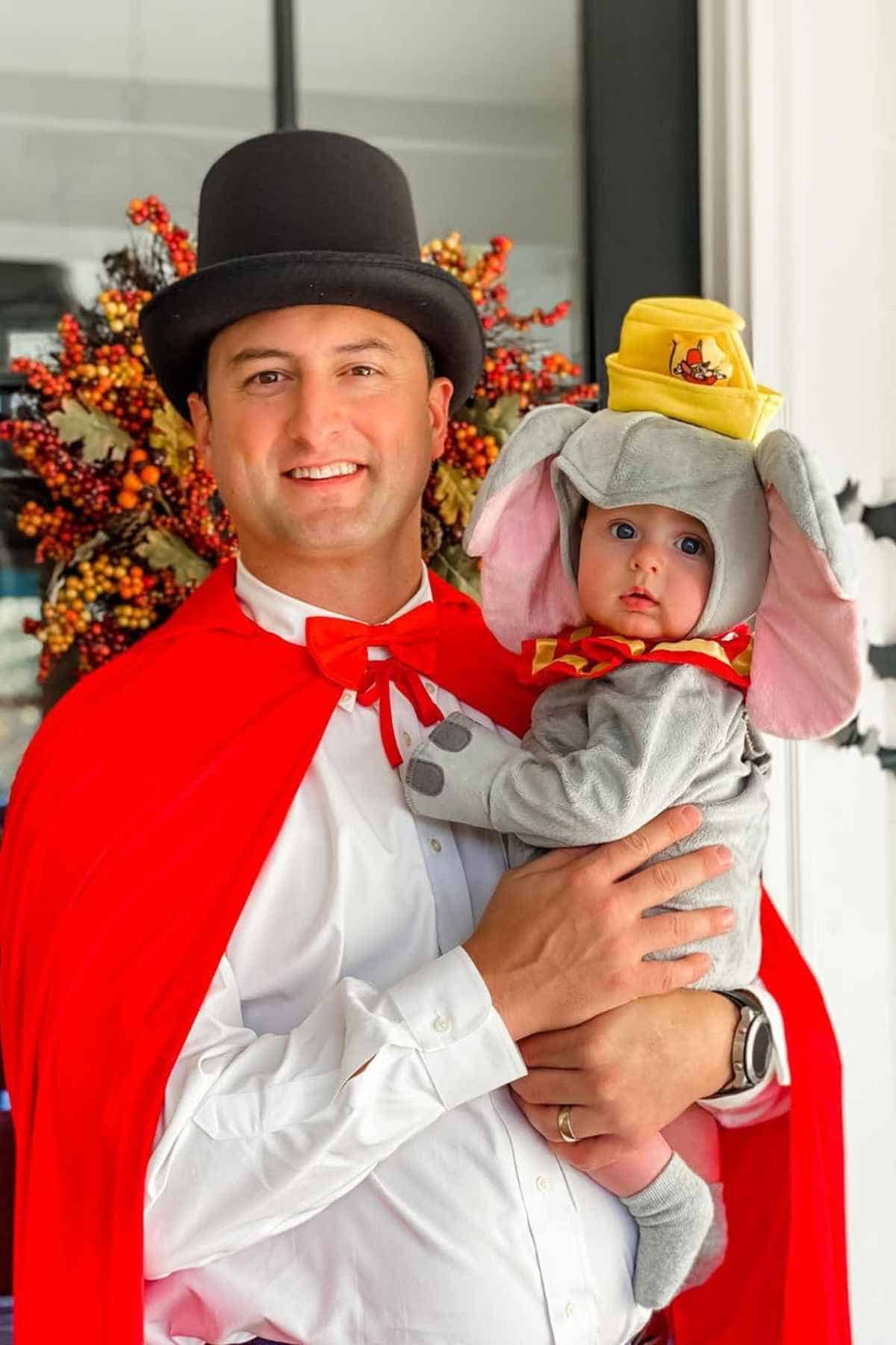 An adult in a top hat and red cape holds a baby dressed in an elephant costume with large ears and a yellow hat.