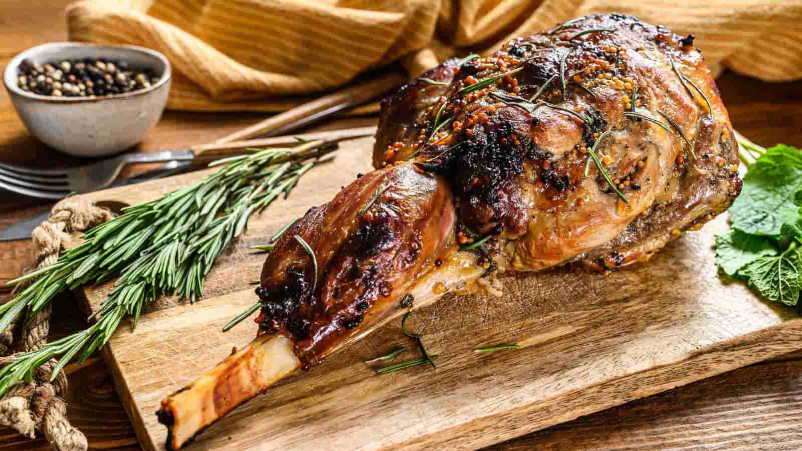 A roasted bone-in leg of lamb garnished with fresh rosemary sits on a wooden cutting board, with a bowl of peppercorns and herbs nearby.