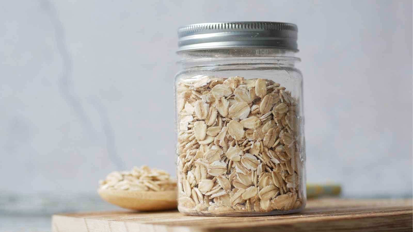 A clear glass jar filled with rolled oats sits on a wooden surface, with a spoonful of oats beside it.