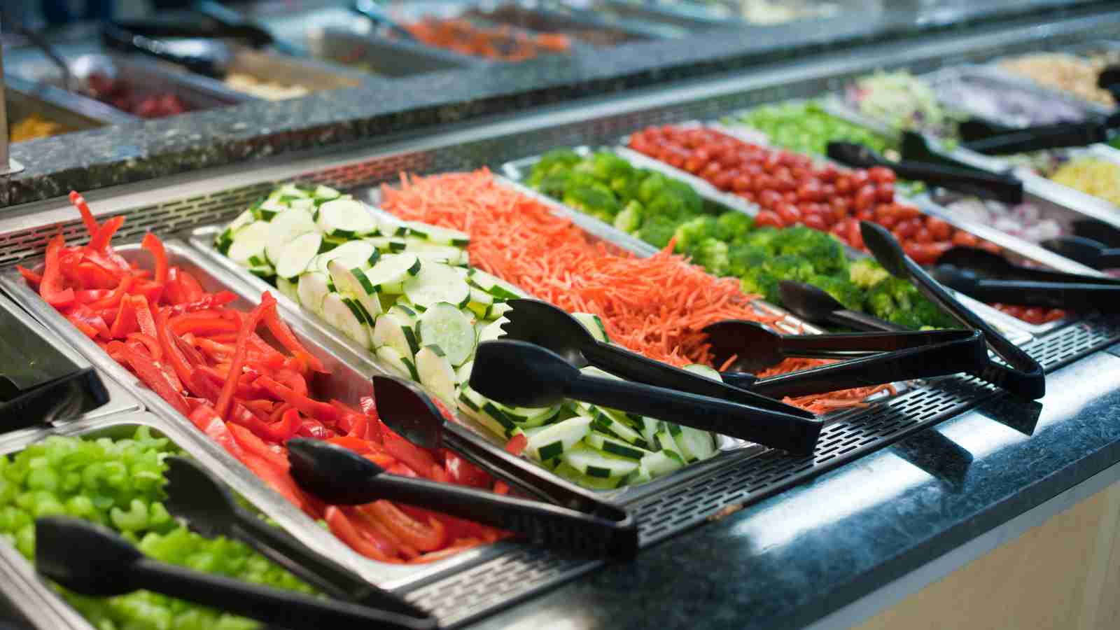 A self-serve salad bar with trays of fresh vegetables, including peppers, cucumbers, carrots, broccoli, and tomatoes, with black serving tongs.