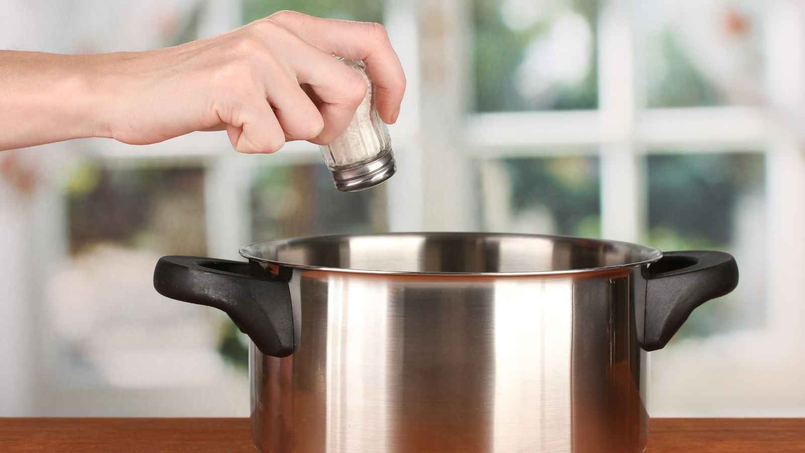 A hand is sprinkling salt from a shaker into a stainless steel pot with black handles on a wooden surface.
