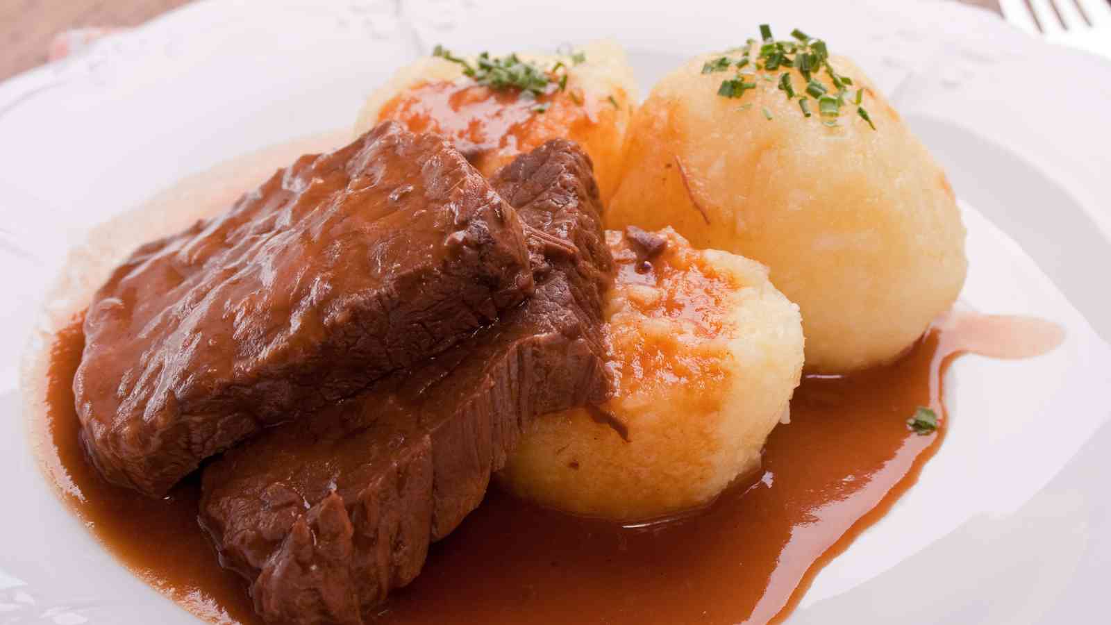 Slices of roast beef with brown gravy served alongside three potato dumplings, garnished with chopped herbs on a white plate.