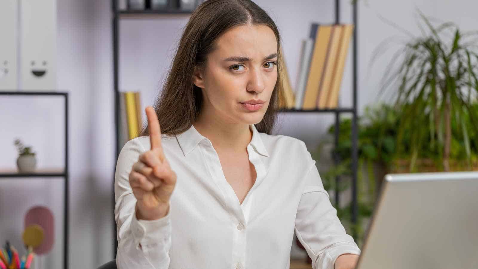 A woman in a white shirt sits at a desk with a laptop, raising her index finger and looking serious, as if emphasizing a point or giving a warning.