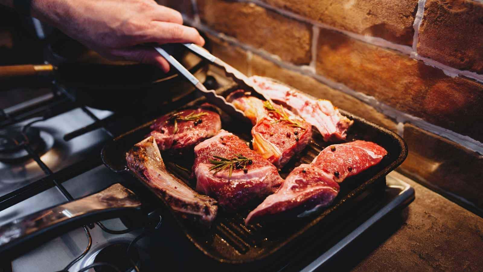 Several raw steaks with herbs are being placed on a grill pan over a stove by a person using metal tongs.