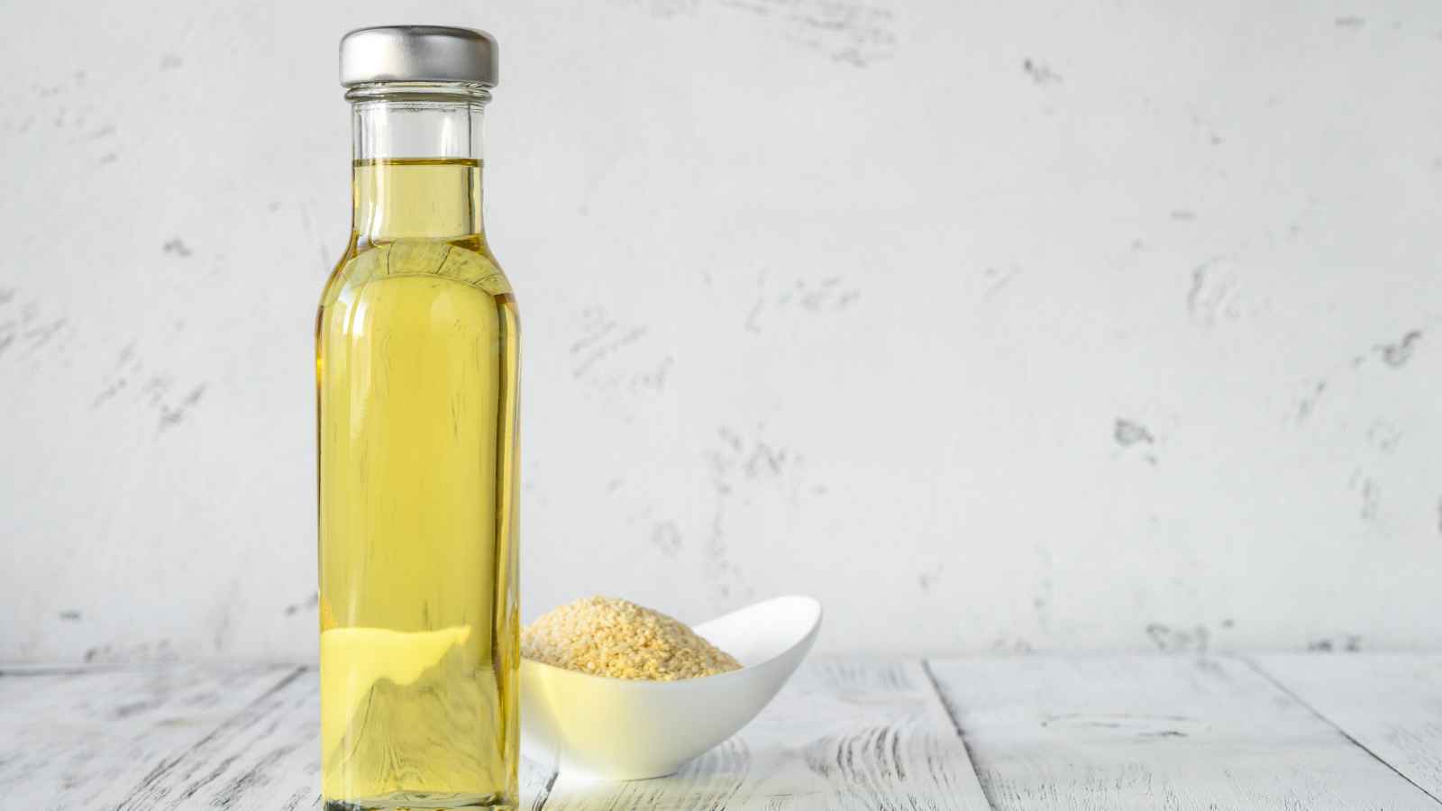 A clear glass bottle filled with yellow oil stands on a white wooden surface next to a white dish containing sesame seeds, against a light textured background.