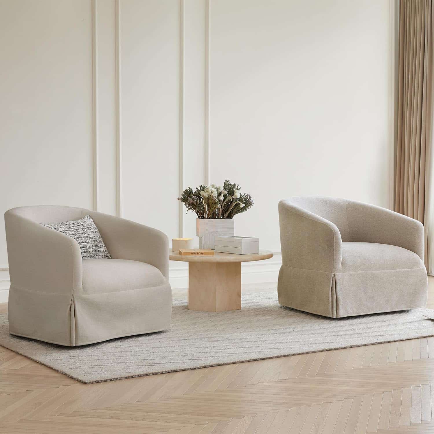 Two beige armchairs with a patterned pillow on one, positioned around a round wooden coffee table with decor, on a rug in a minimalist, light-filled room.