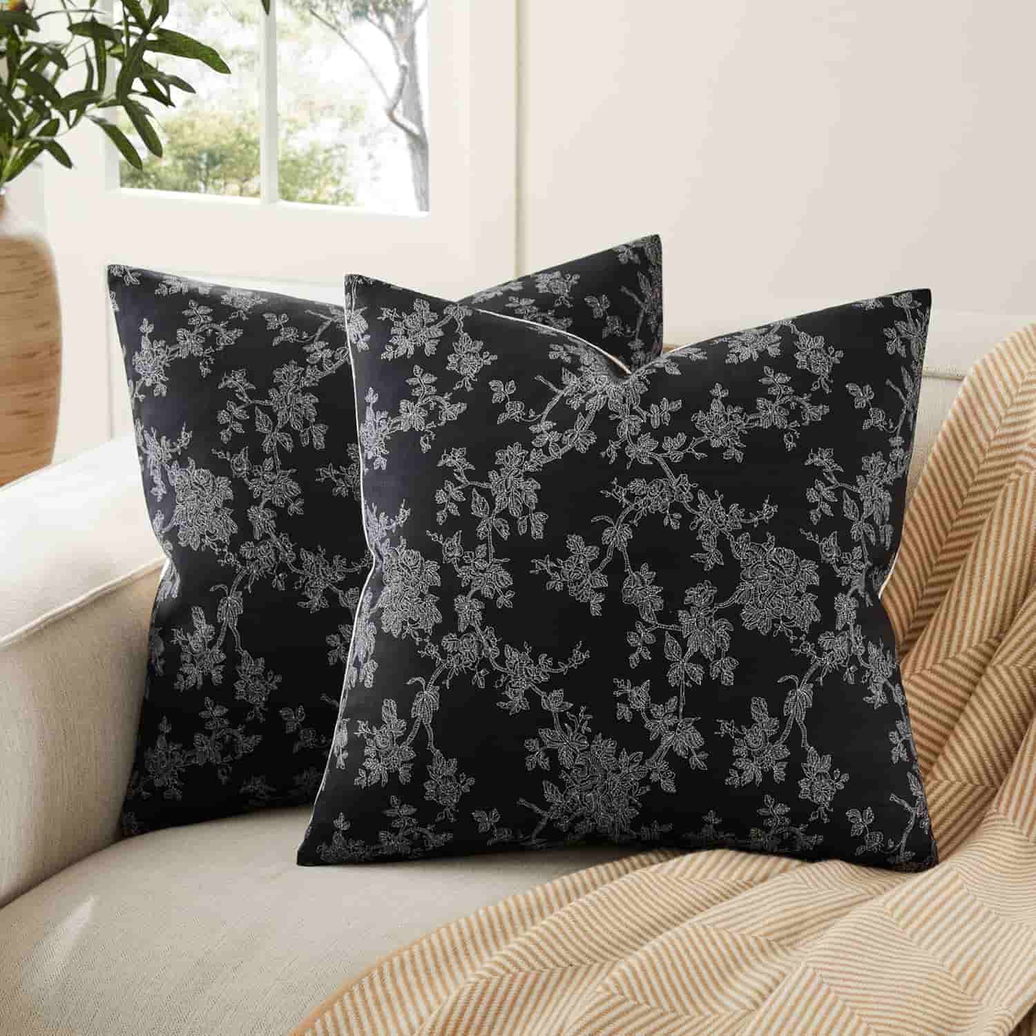 Two black throw pillows with white floral patterns are placed on a beige couch next to a striped tan blanket, with a window in the background.