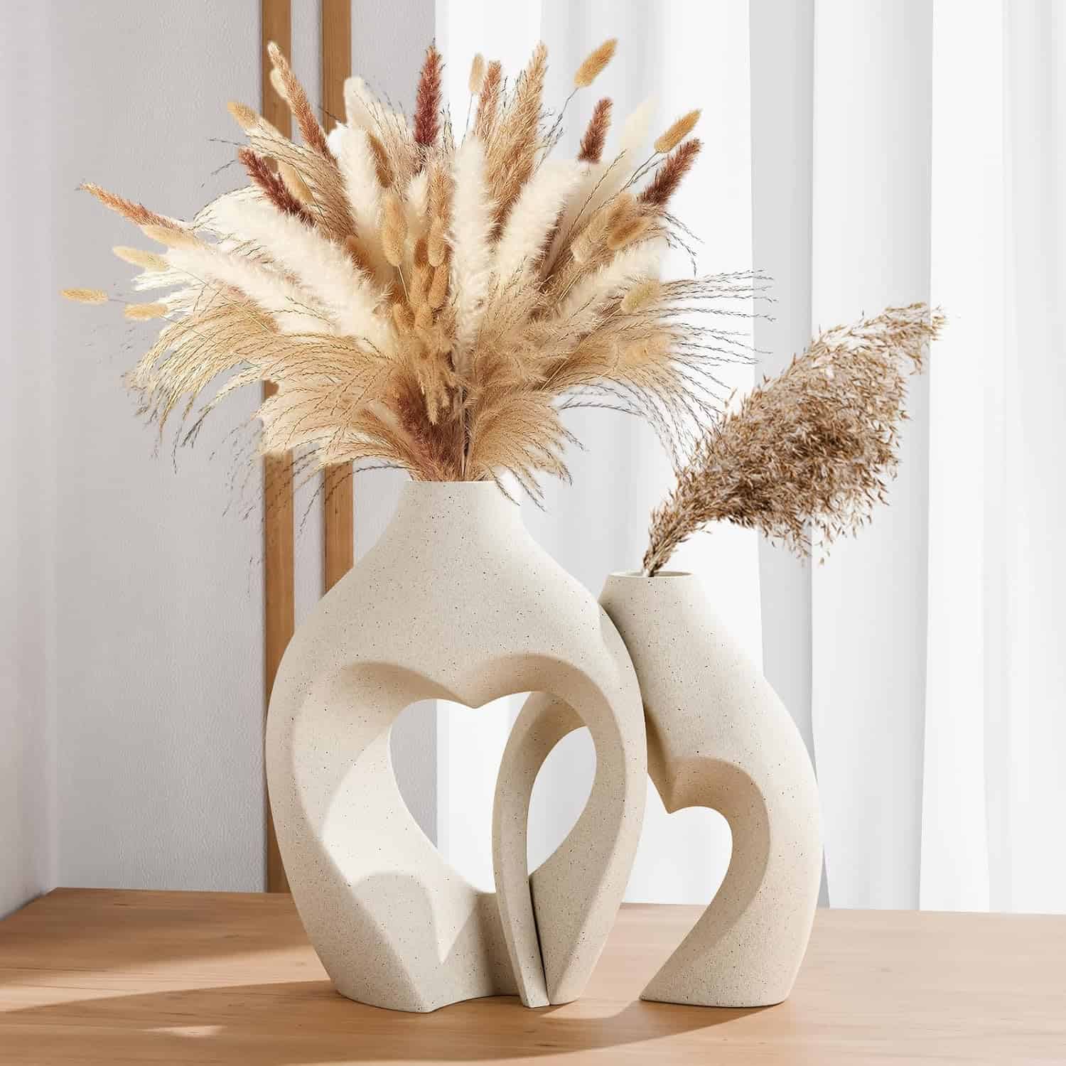 Two modern ceramic vases with heart-shaped cutouts sit on a wooden surface, each holding dried pampas grass and other beige foliage. White curtains hang in the background.