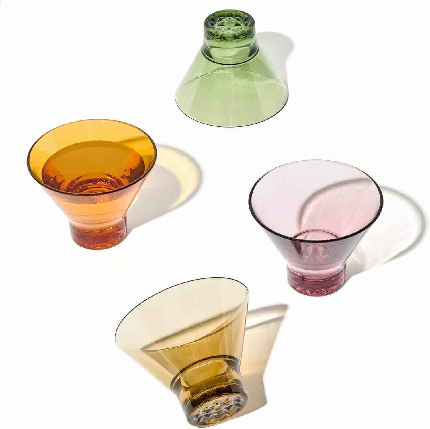 Four colored glass tumblers—orange, green, pink, and yellow—are arranged on a white background, with two upright and two tilted on their sides.