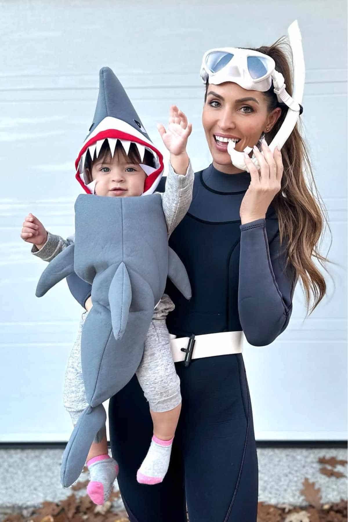 Woman in scuba gear holds a baby dressed in a shark costume; both are smiling and posing for the camera in front of a light-colored wall.