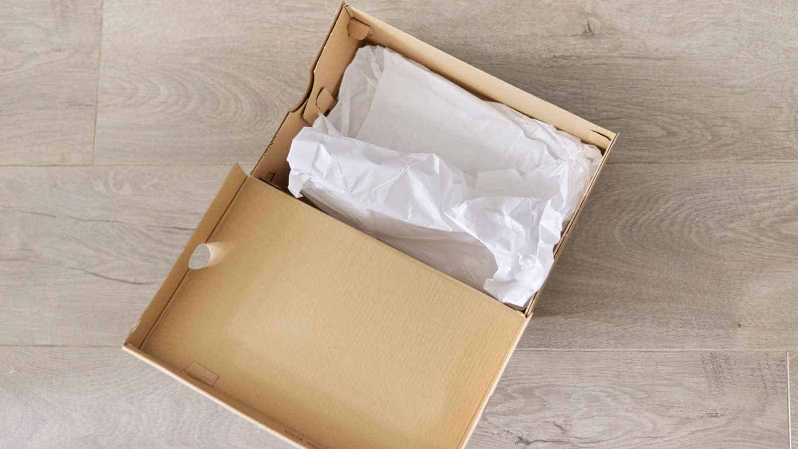 An open cardboard shoe box with white tissue paper inside, placed on a light-colored wooden floor.