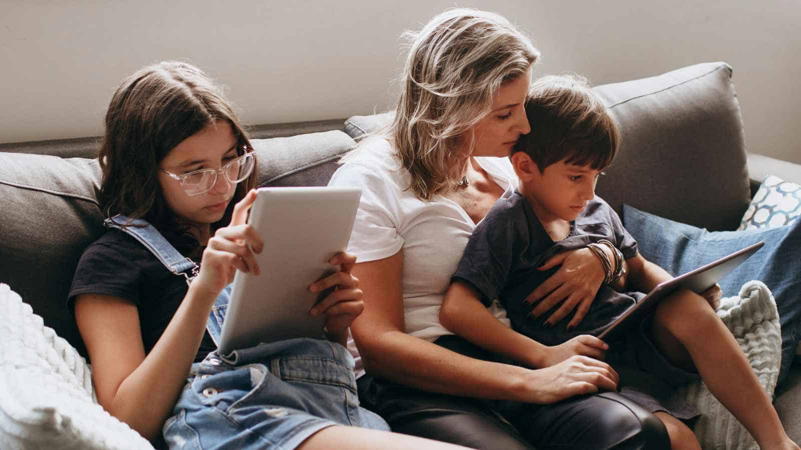 A woman sits on a couch with a young boy in her lap and a girl beside her; both children are using tablets.