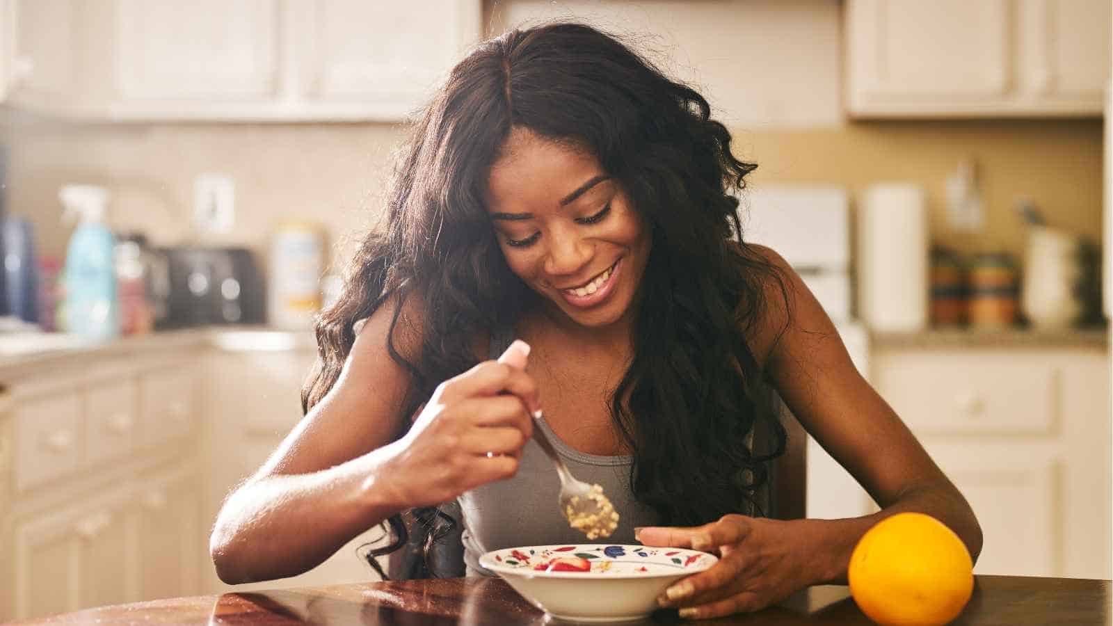 A woman sits at a kitchen table, smiling while eating from a bowl with a spoon. An orange rests on the table beside her.