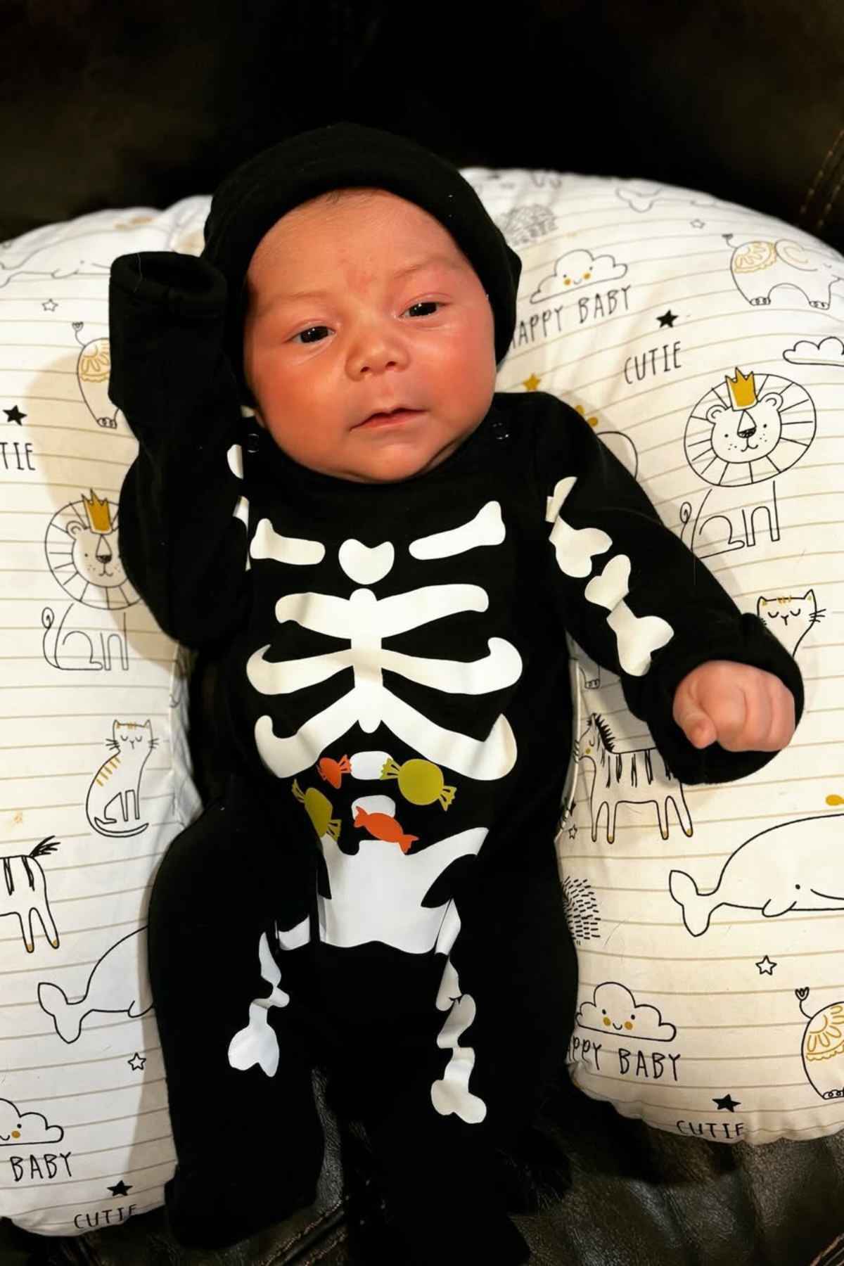 A newborn baby dressed in a black skeleton costume lies on a pillow decorated with cartoon lions and hippos.