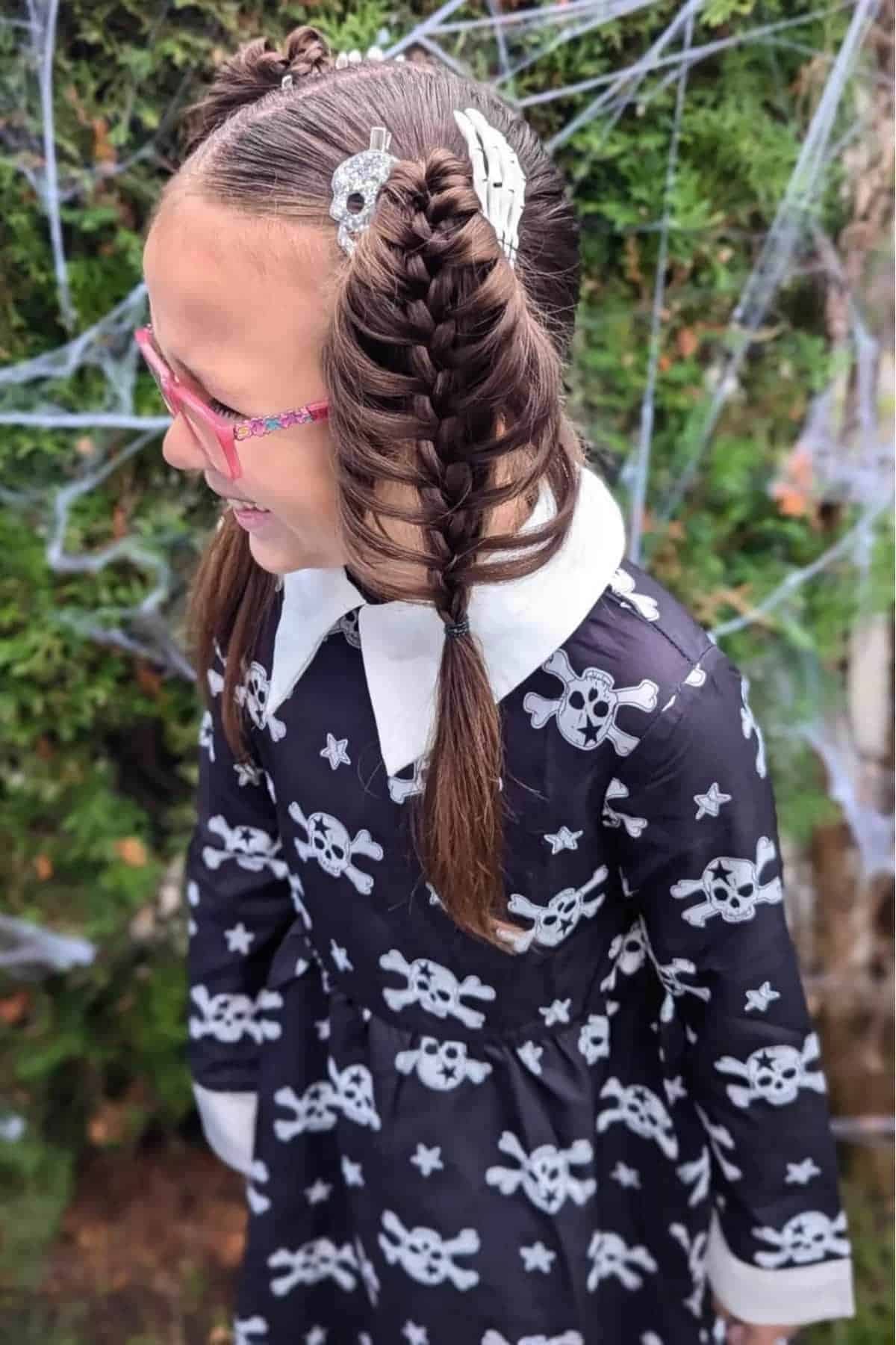 A child wearing a black dress with white skull patterns and pink glasses, with fishtail braids decorated with skull hair clips, standing outdoors near fake spider webs.
