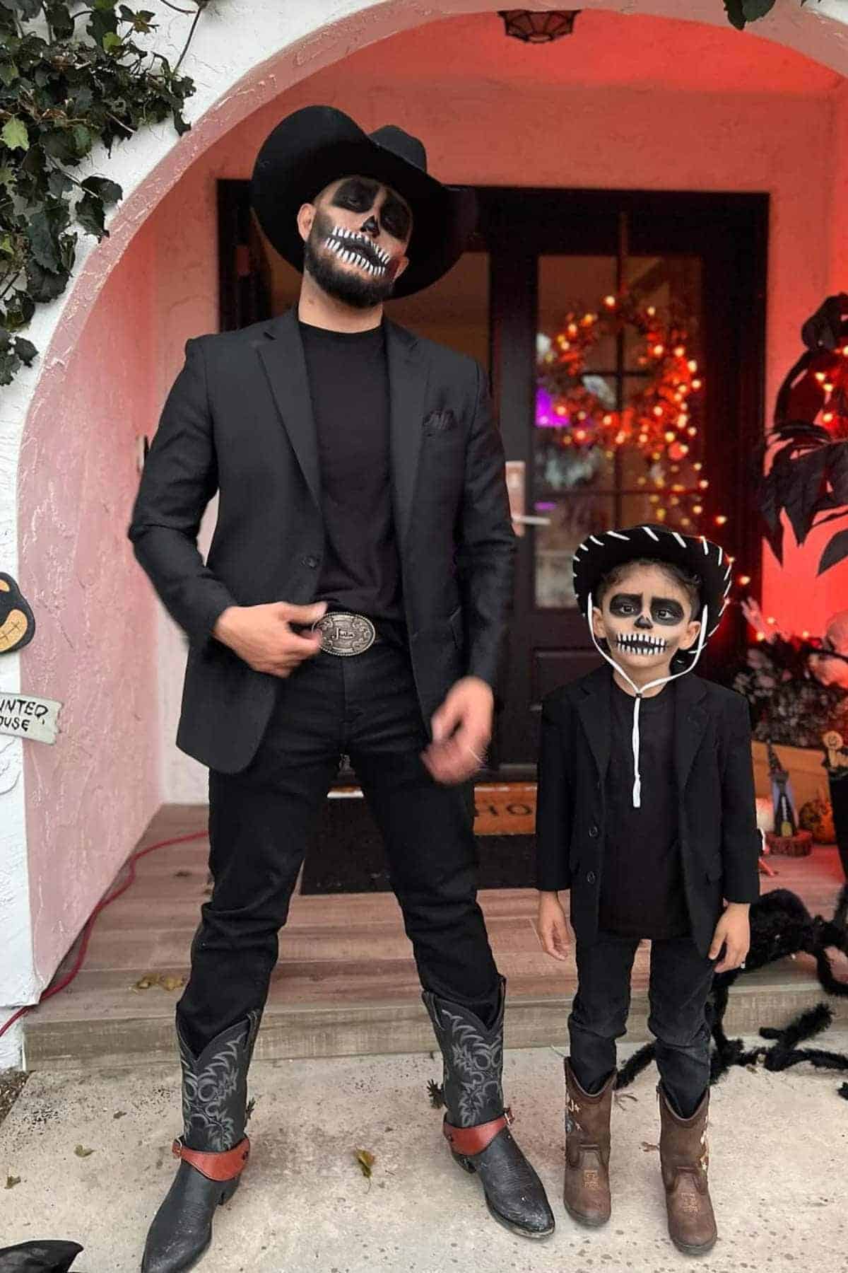 An adult and a child in matching black outfits and cowboy hats, with painted skull faces, stand in front of a decorated doorway.