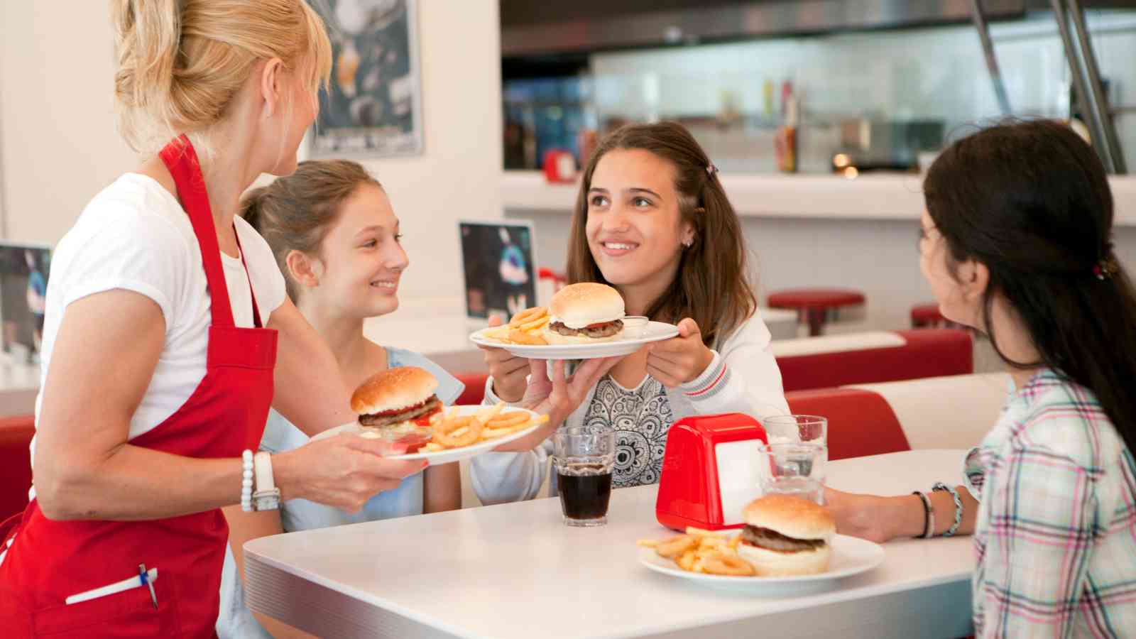 A server in a red apron hands plates of burgers and fries to three girls sitting at a diner table.