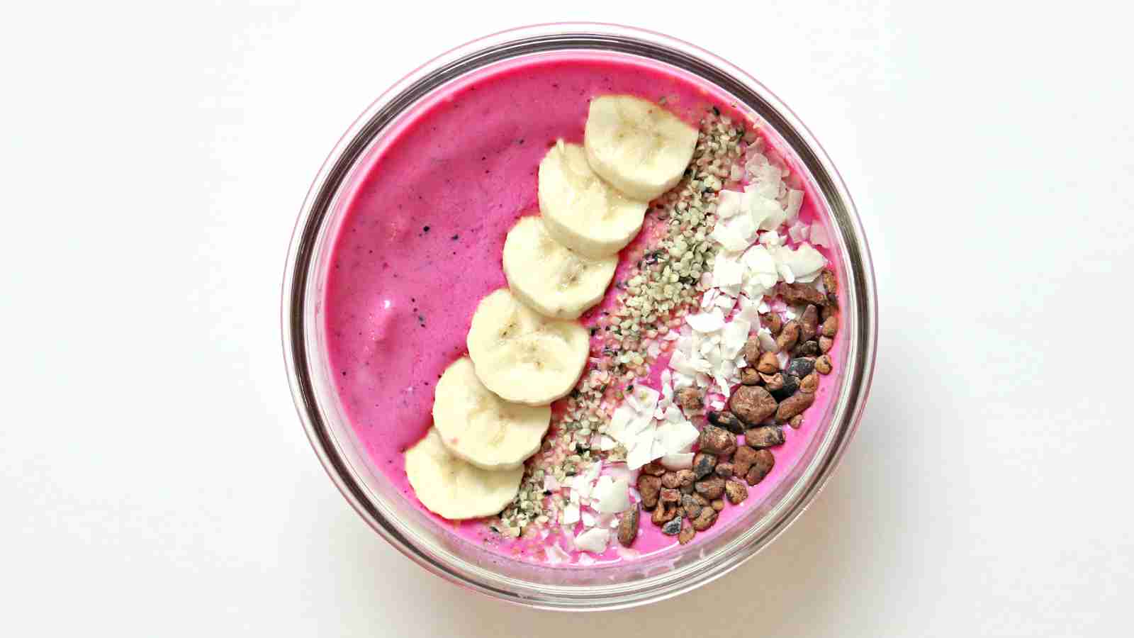 A smoothie bowl with bright pink base, topped with banana slices, hemp seeds, shredded coconut, and cacao nibs, arranged in neat rows.
