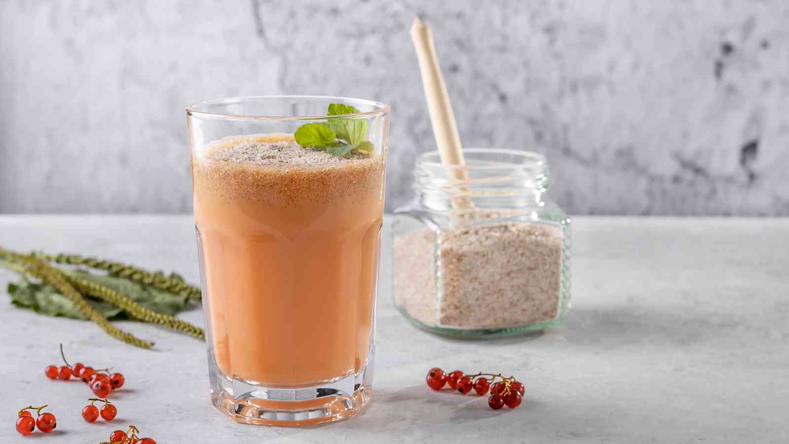 A glass of orange drink garnished with mint is on a table next to a jar filled with powder and a wooden spoon, with red berries and green leaves nearby.