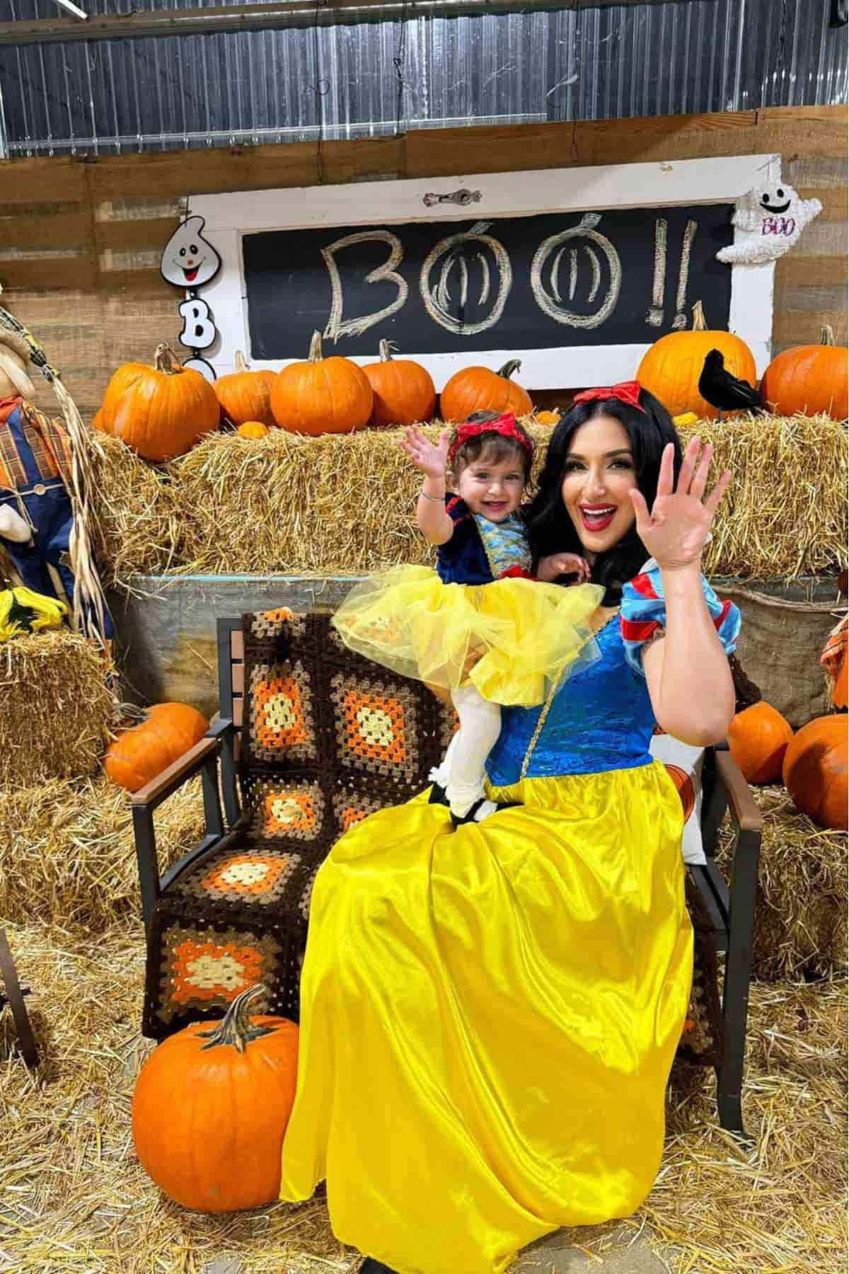 Two people dressed as Snow White sit on a bench surrounded by pumpkins and hay bales, waving and smiling at the camera in a festive Halloween setting.