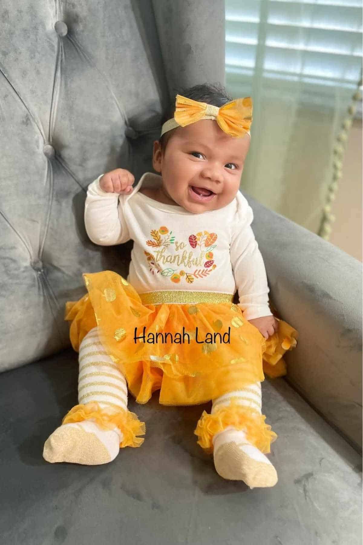 Baby wearing a yellow bow headband, " so thankful" shirt, yellow skirt, and striped leggings, sitting and smiling on a gray chair.