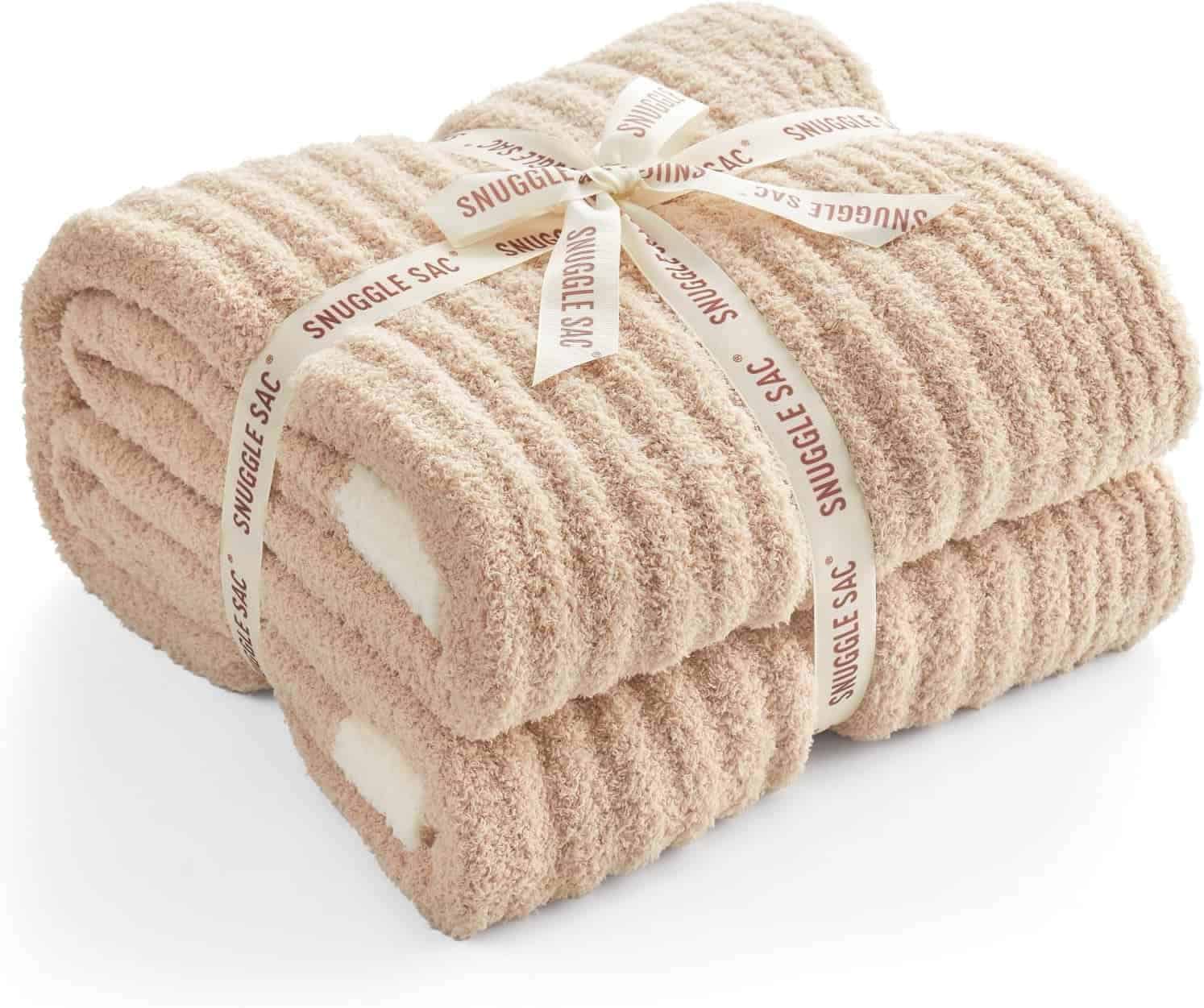 Two beige, ribbed fleece blankets are folded and stacked, tied together with a cream ribbon that reads "SNUGGLE SAC" in brown text.