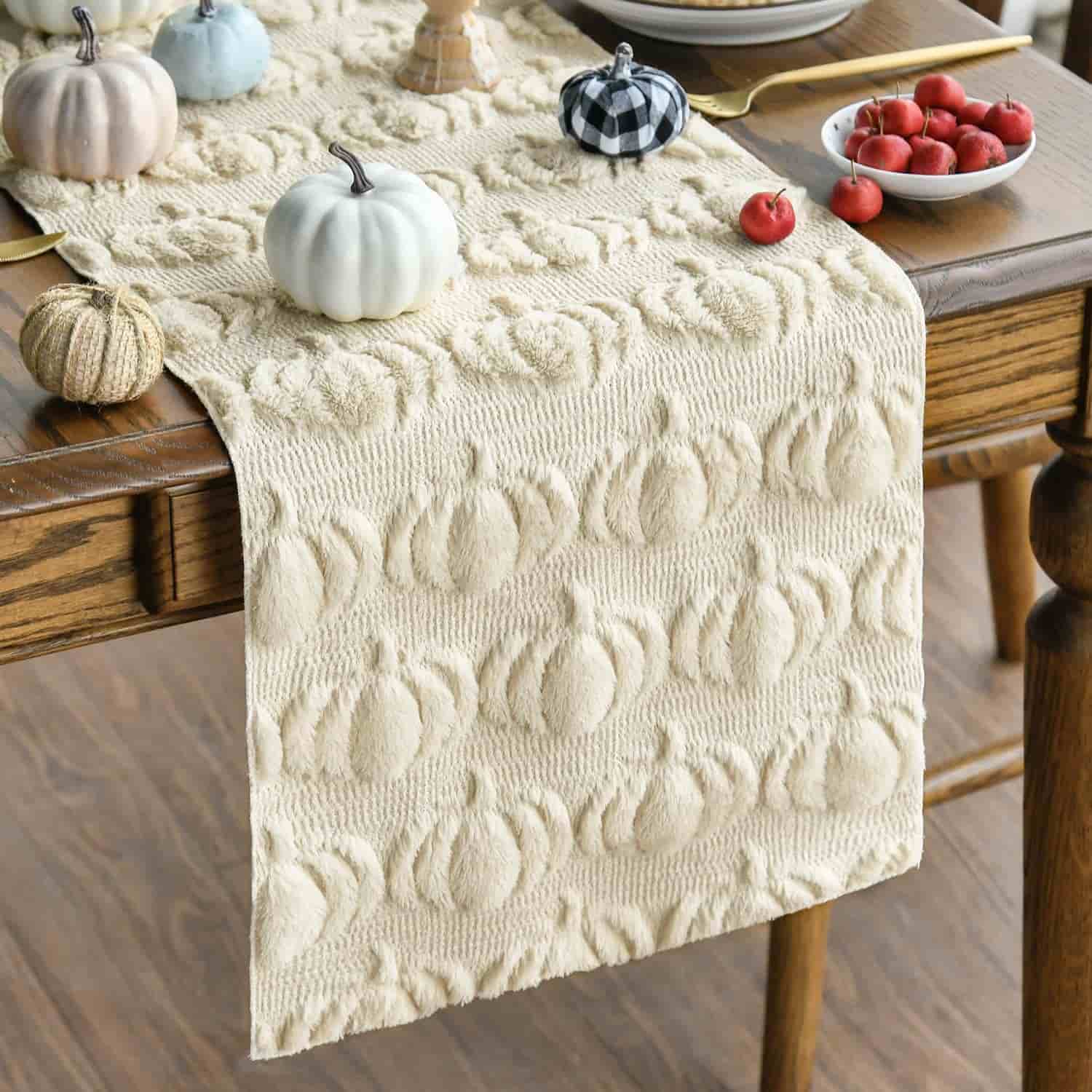 A beige textured table runner with a pumpkin pattern on a wooden table, decorated with small pumpkins and bowls of fruit.