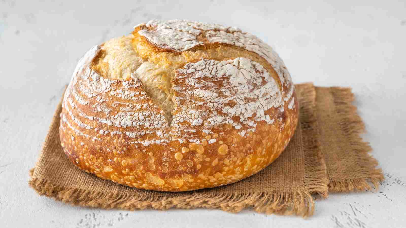 A round loaf of rustic sourdough bread with a cracked, flour-dusted crust sits on a piece of burlap fabric against a light background.