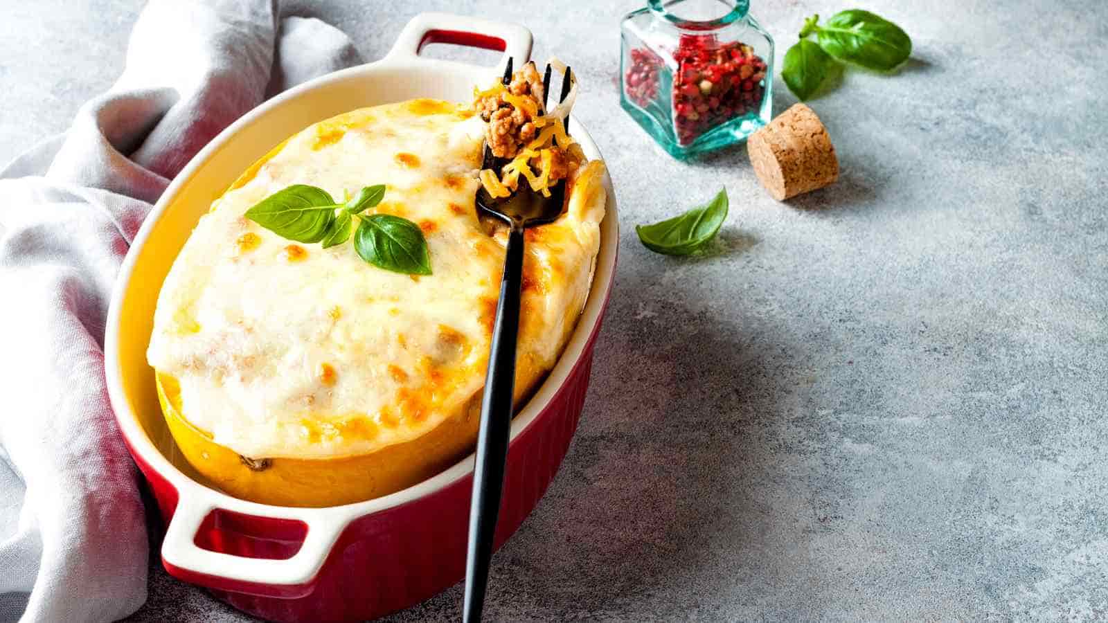 A baked casserole topped with melted cheese and basil leaves sits in a red dish, with a serving spoonful lifted out. A jar of peppercorns and a cork are nearby.