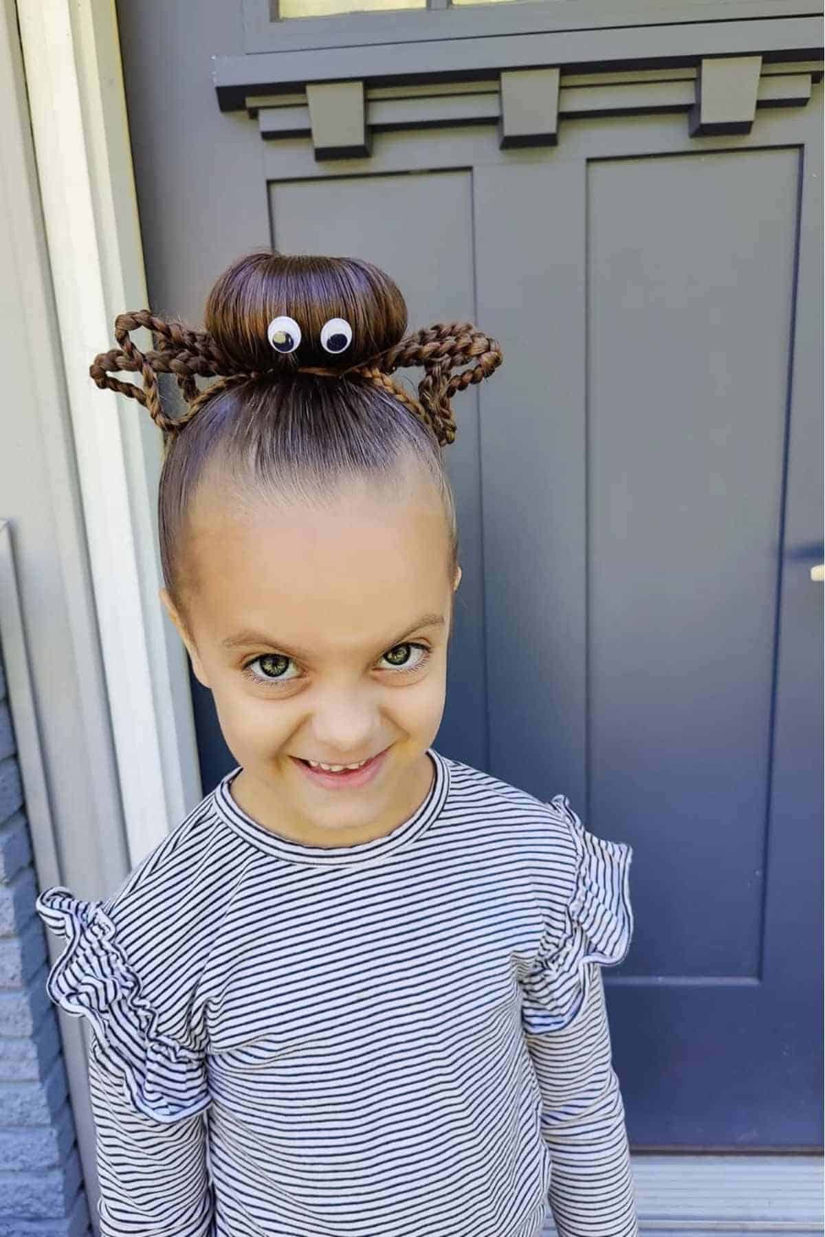 A young girl with a high bun styled as a spider using braided hair legs and googly eyes, standing in front of a gray door, wearing a striped shirt with ruffled sleeves.