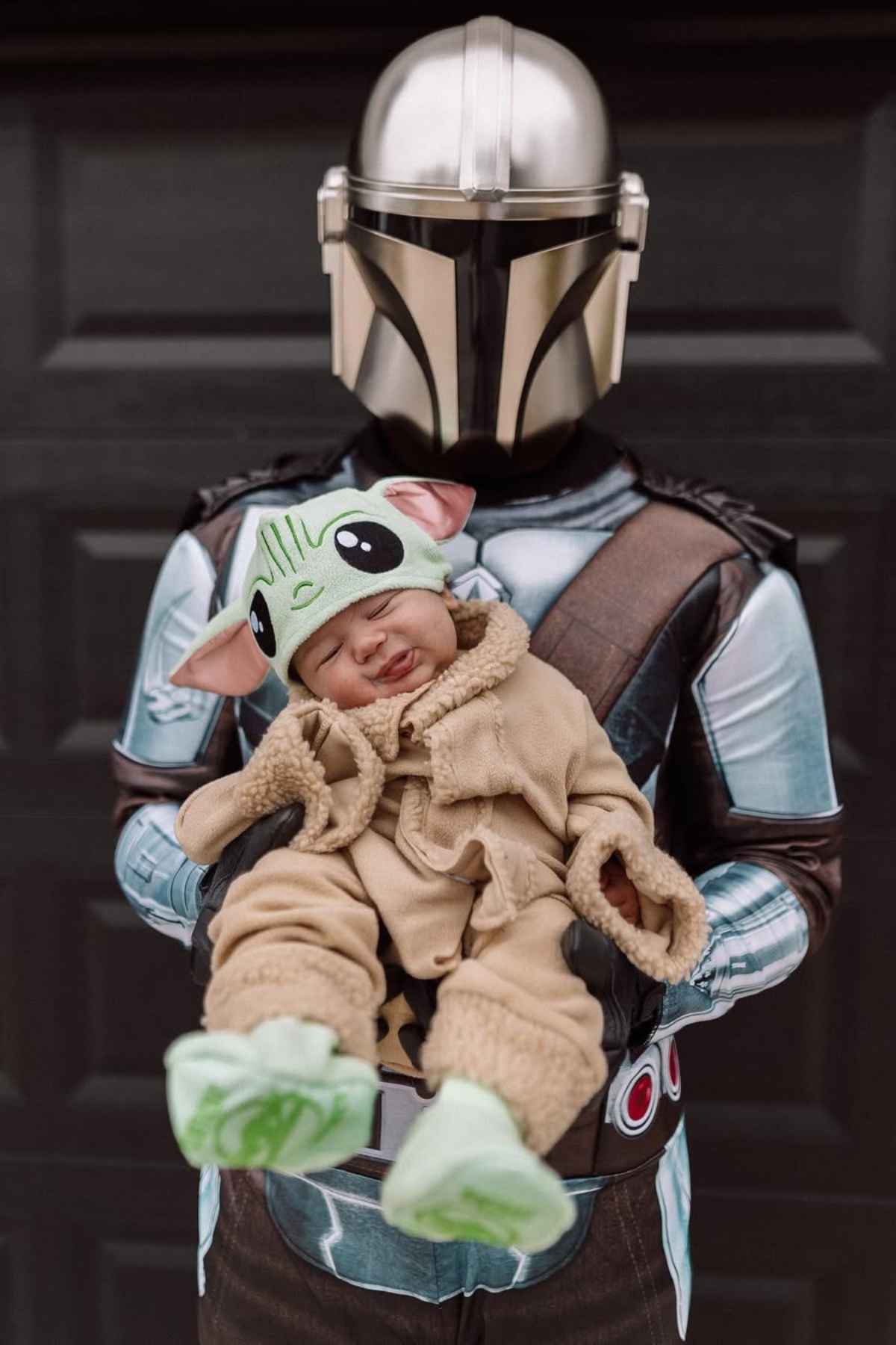 An adult dressed as the Mandalorian holds a baby wearing a Grogu (Baby Yoda) costume in front of a dark background.