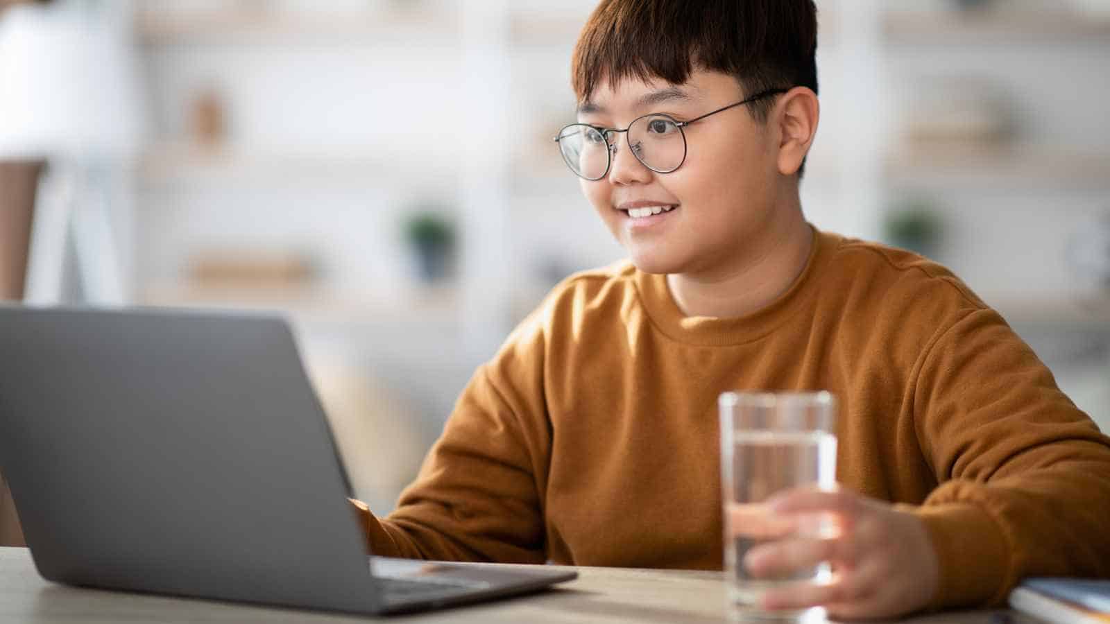 A person wearing glasses and a brown sweater uses a laptop at a desk, holding a glass of water. The background is softly blurred.