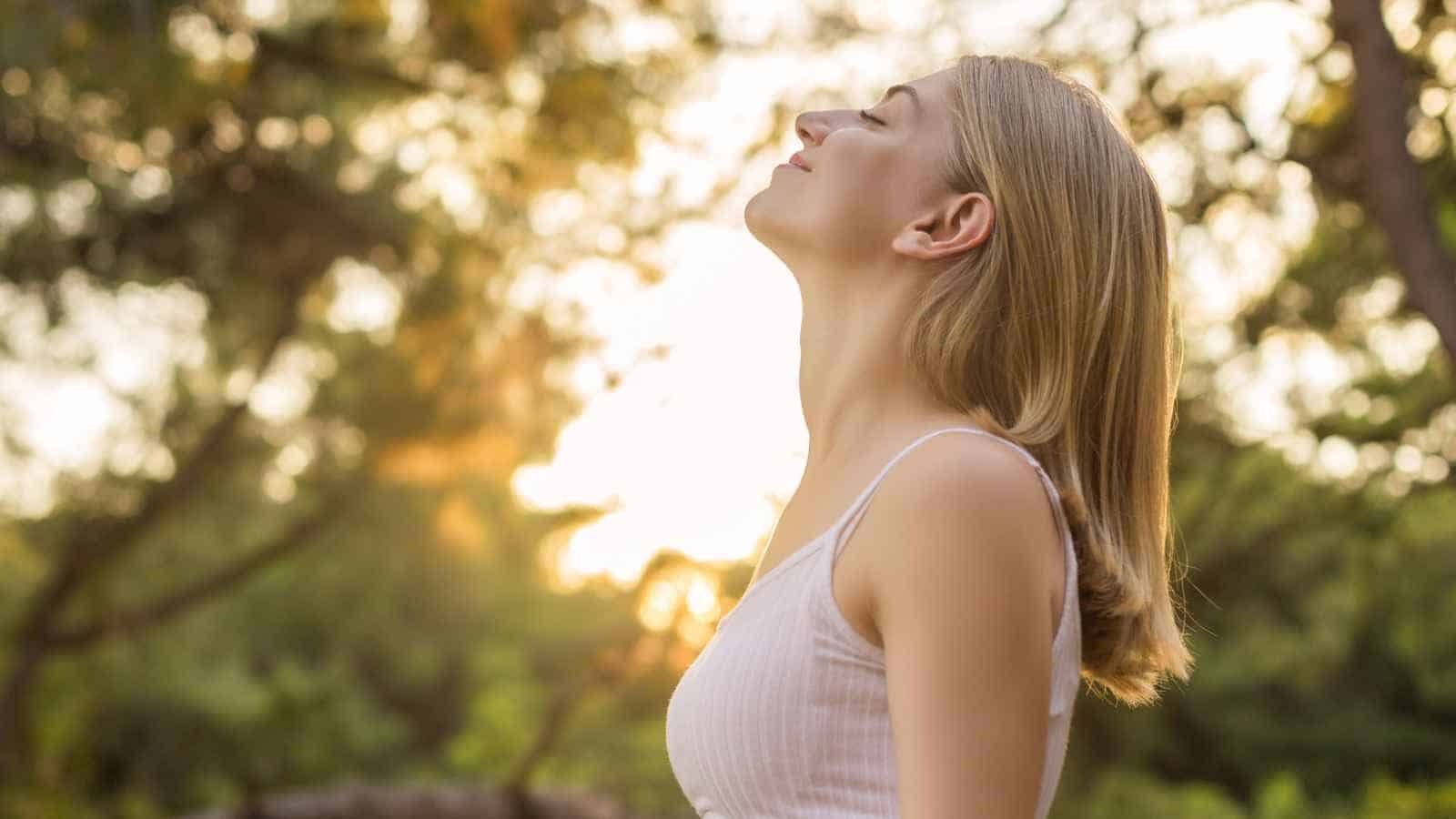 A woman with blonde hair stands outdoors with her eyes closed and head slightly tilted back, appearing relaxed. Sunlight and greenery are visible in the background.
