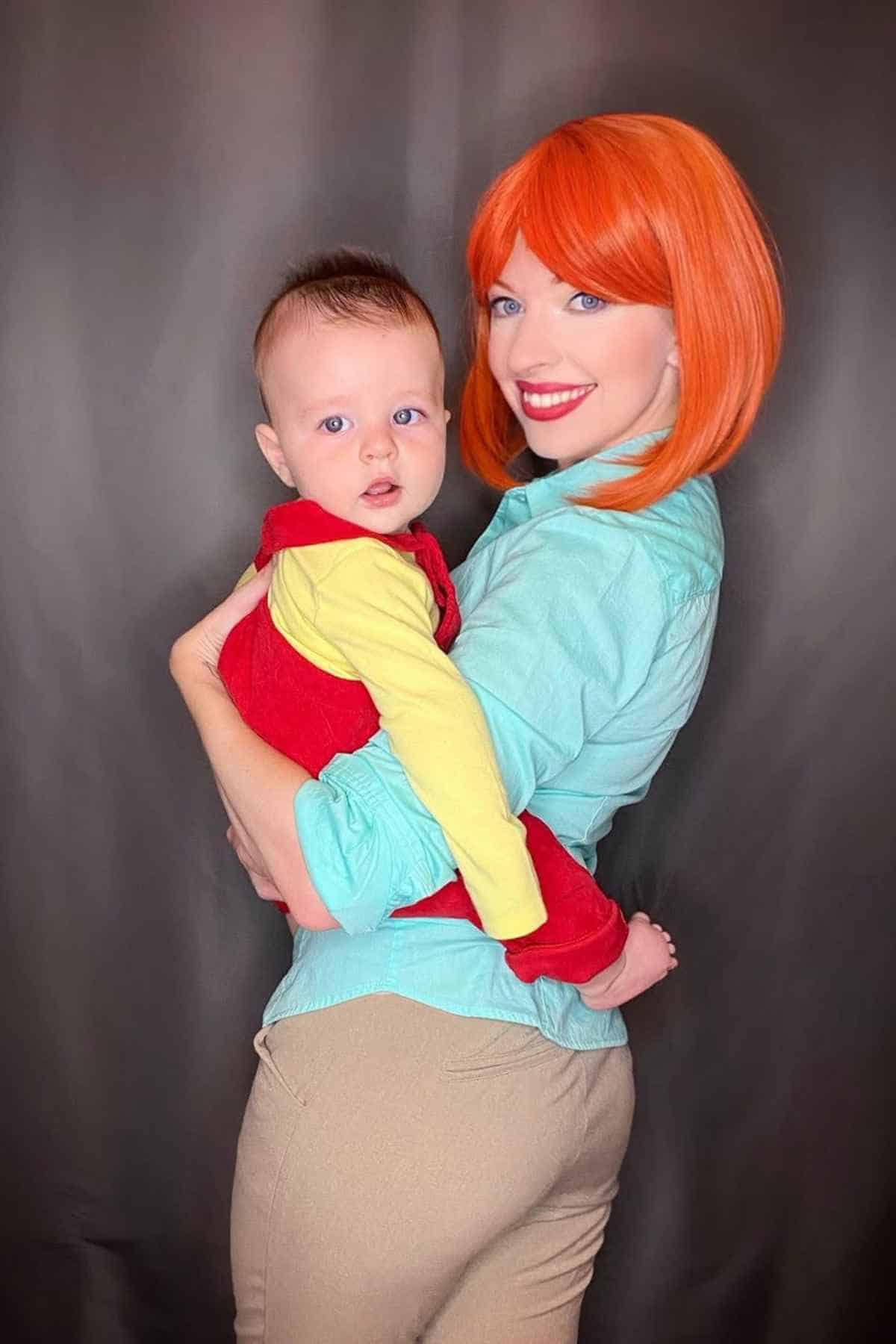 A woman with a bright orange bob wig holds a baby dressed in a red and yellow outfit, both facing the camera against a plain dark background.
