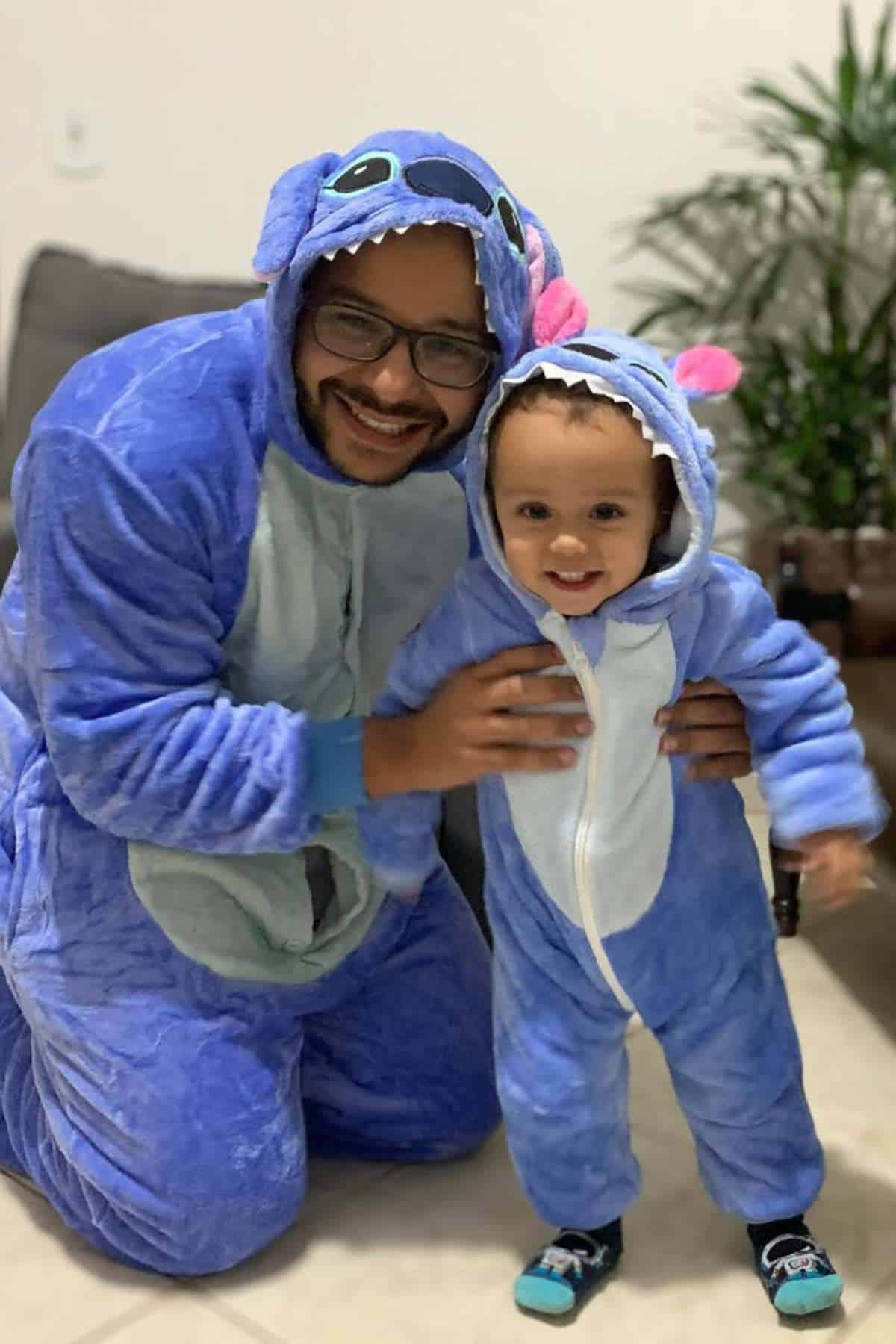 An adult and a child, both wearing matching blue Stitch onesies, smile at the camera indoors.