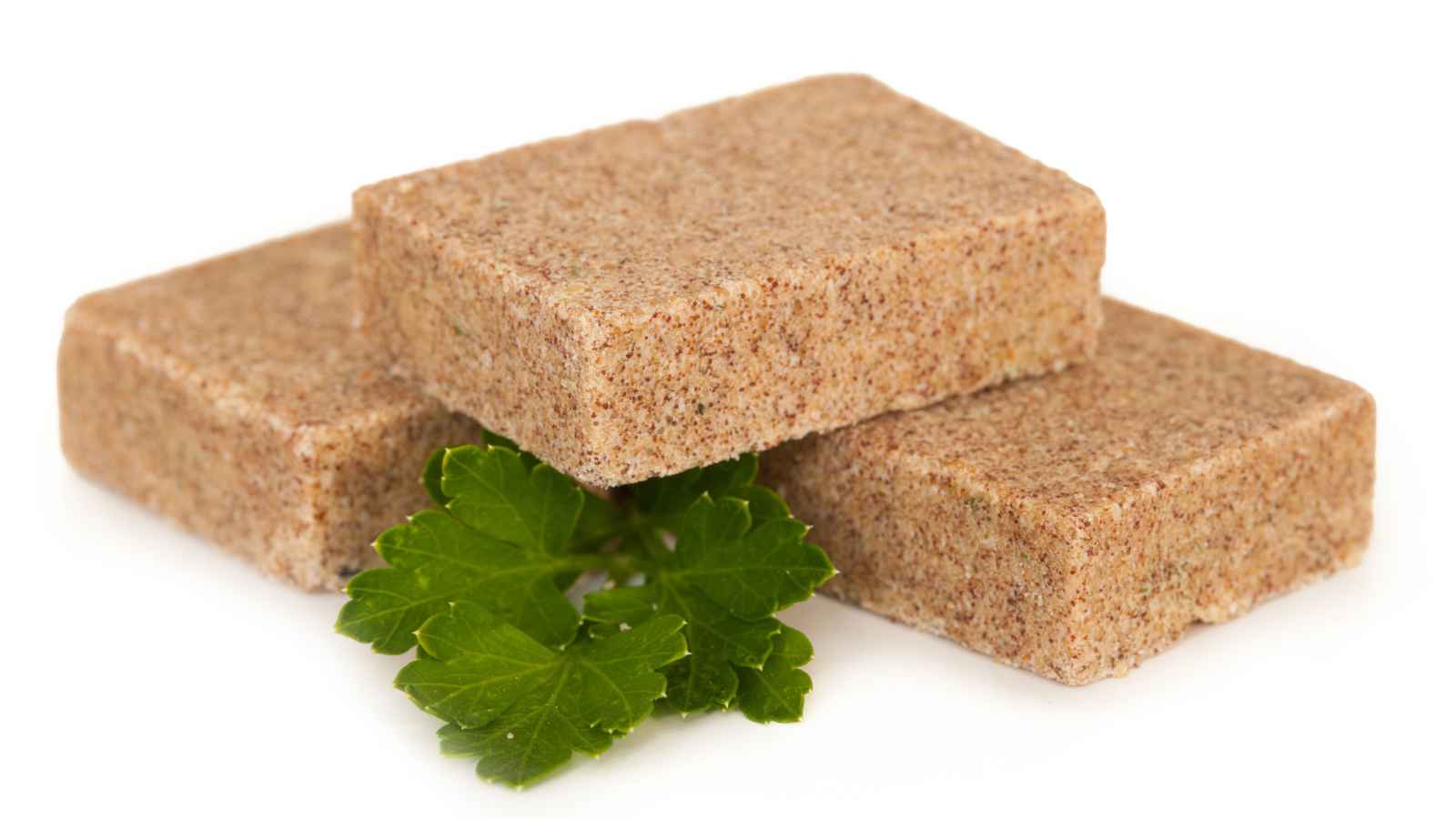 Three rectangular brown bouillon cubes stacked with a sprig of parsley on a white background.