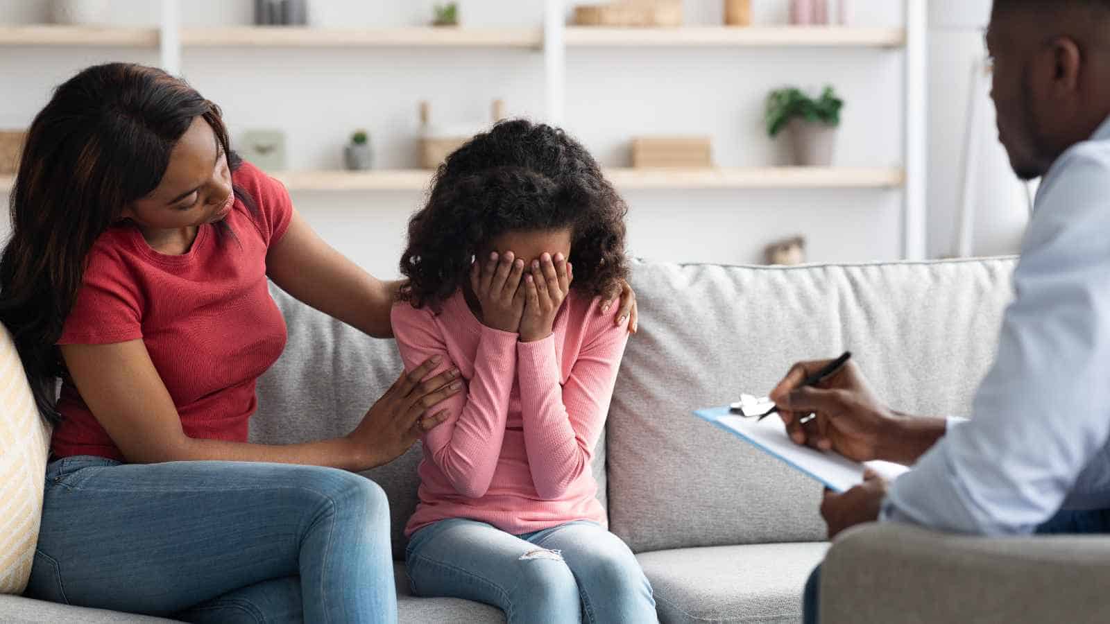 A woman comforts a girl who is covering her face and crying while a man with a clipboard takes notes during a counseling session.