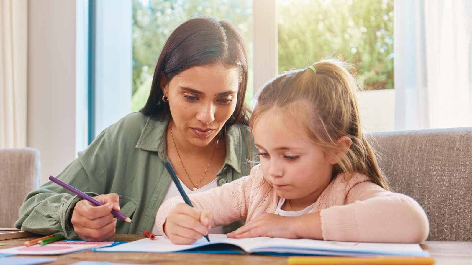 An adult and a young girl sit at a table, both writing or drawing on paper, with colored pencils and a window in the background.